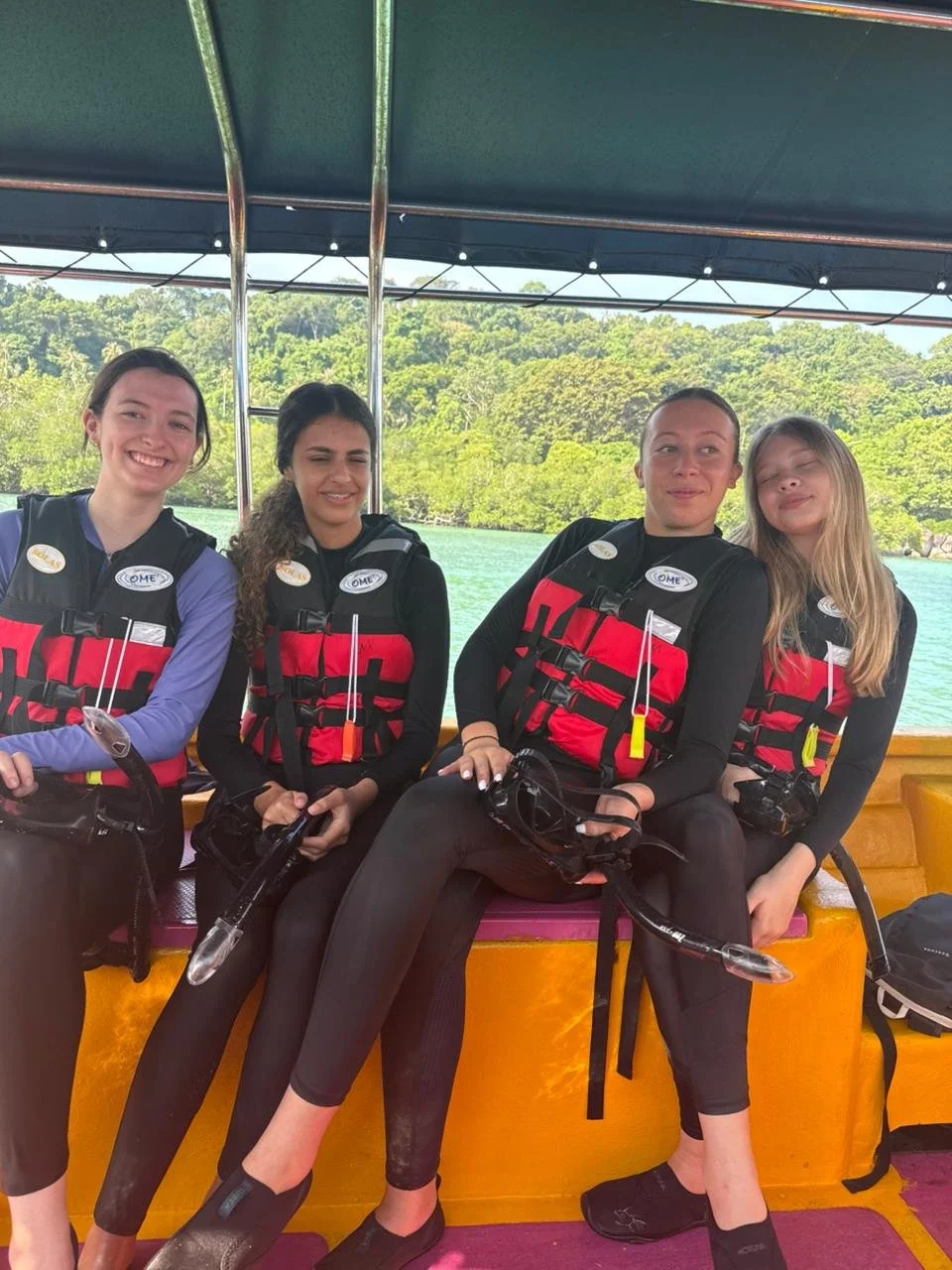 Four women sitting on a boat, wearing life jackets, with water and greenery in the background.