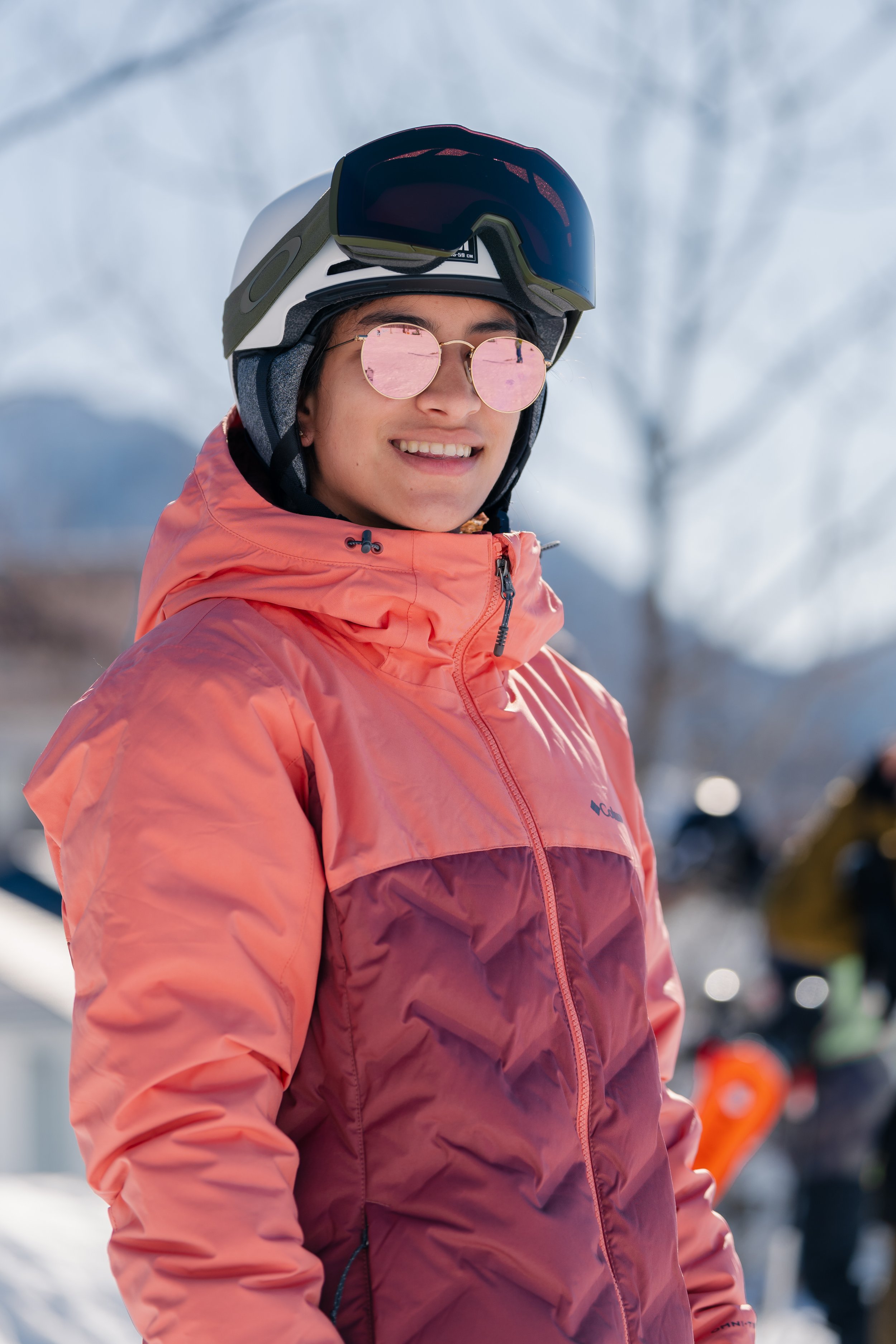 Close-up of a smiling woman wearing a white ski helmet with goggles and pink mirrored sunglasses, dressed in a pink and purple winter jacket, snowy background with blurred trees and snow-covered ground.
