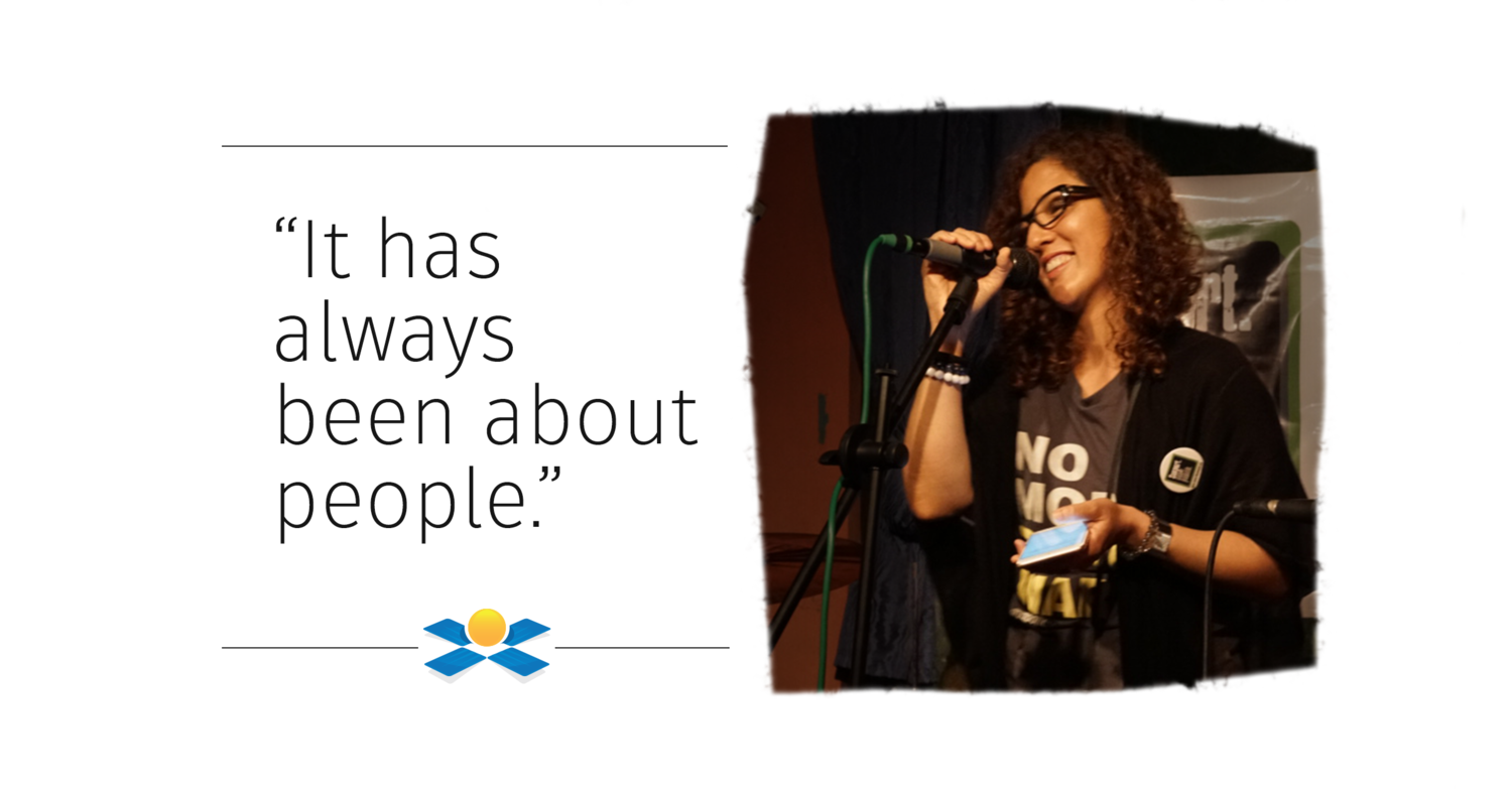 A woman with curly hair, glasses, and a black T-shirt smiling and speaking into a microphone on stage, holding a phone in her hand.