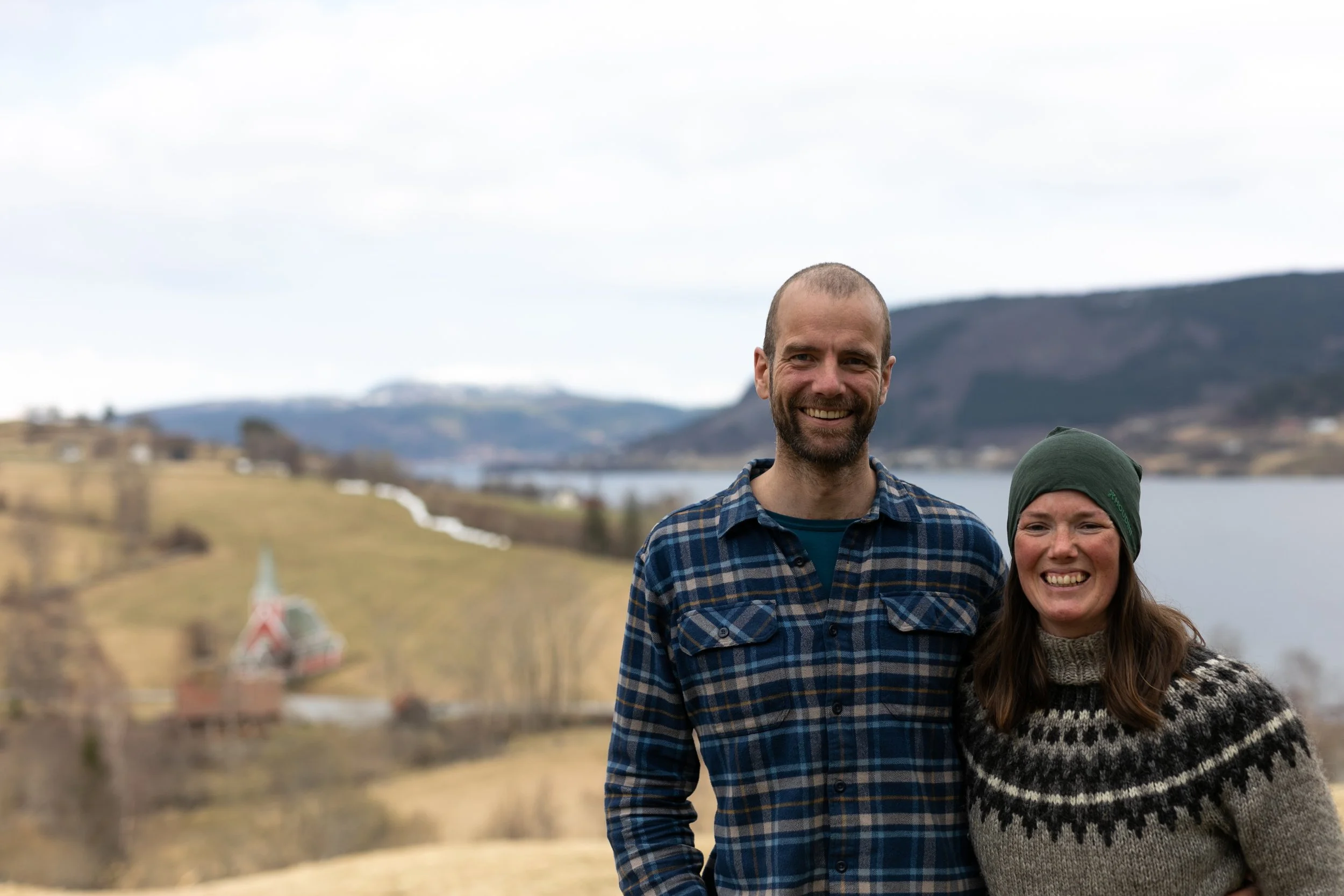 To personer smiler foran et landskap med vann, åser og spredte bygninger.