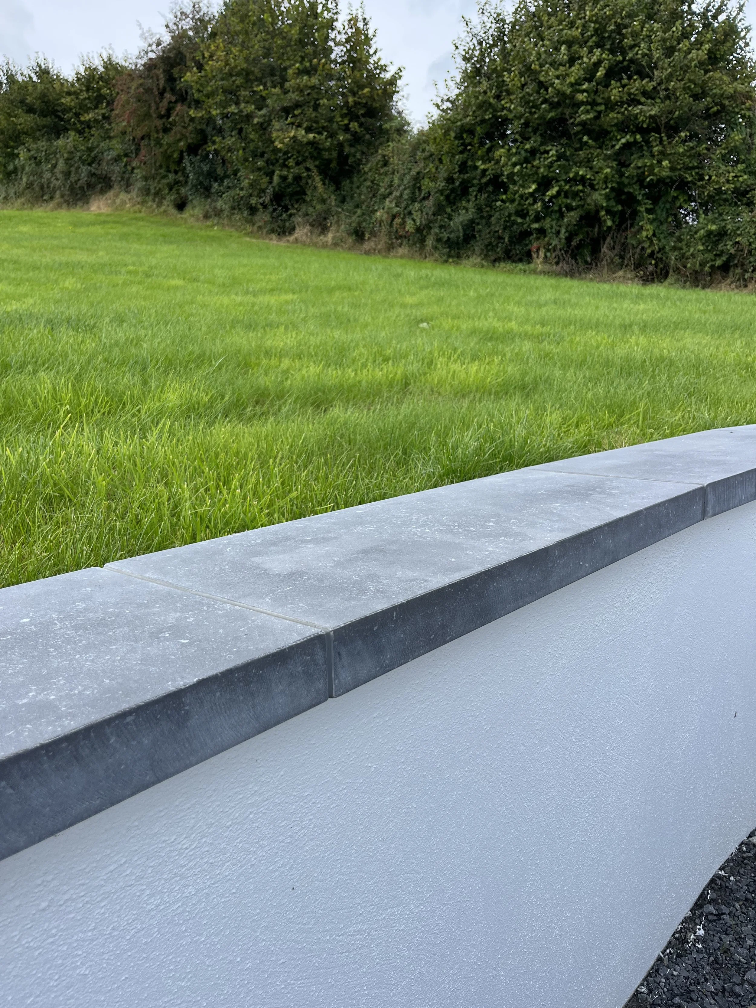 A concrete sidewalk with a dark gray stone border and a grassy hill with trees in the background.
