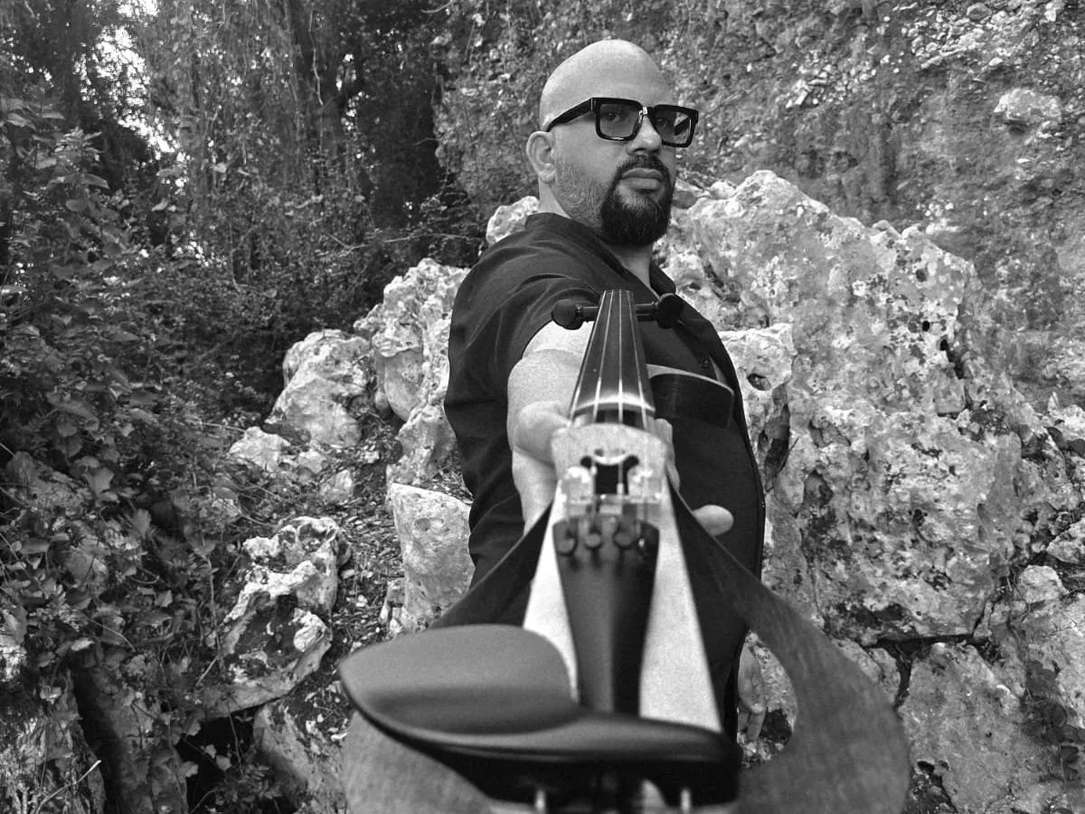 A man taking a selfie outdoors, pointing a rifle toward the camera, with a rocky and wooded background.