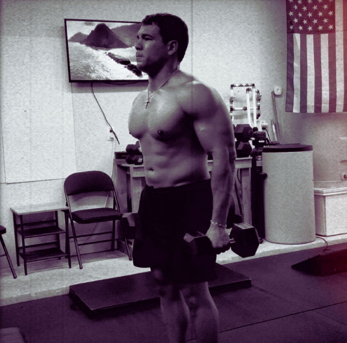 A shirtless man holding a dumbbell in a gym setting with an American flag on the wall.