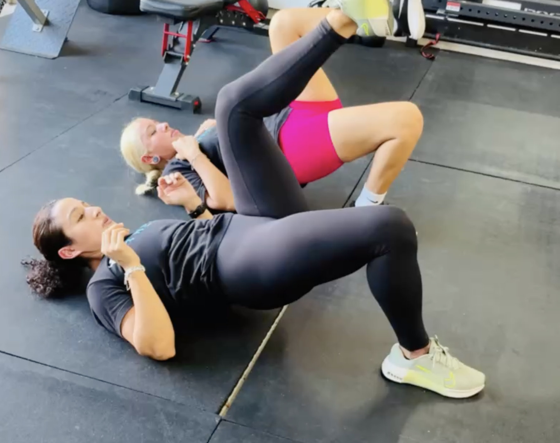 Two women exercising on gym mats, performing glute bridge exercises, with one woman in teal leggings and a black t-shirt and the other in pink shorts and a black t-shirt.