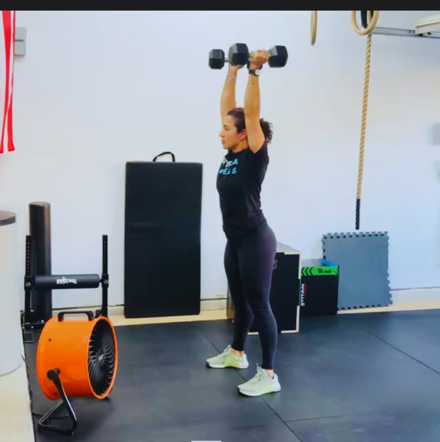 A woman lifting dumbbells overhead during a workout in a gym.