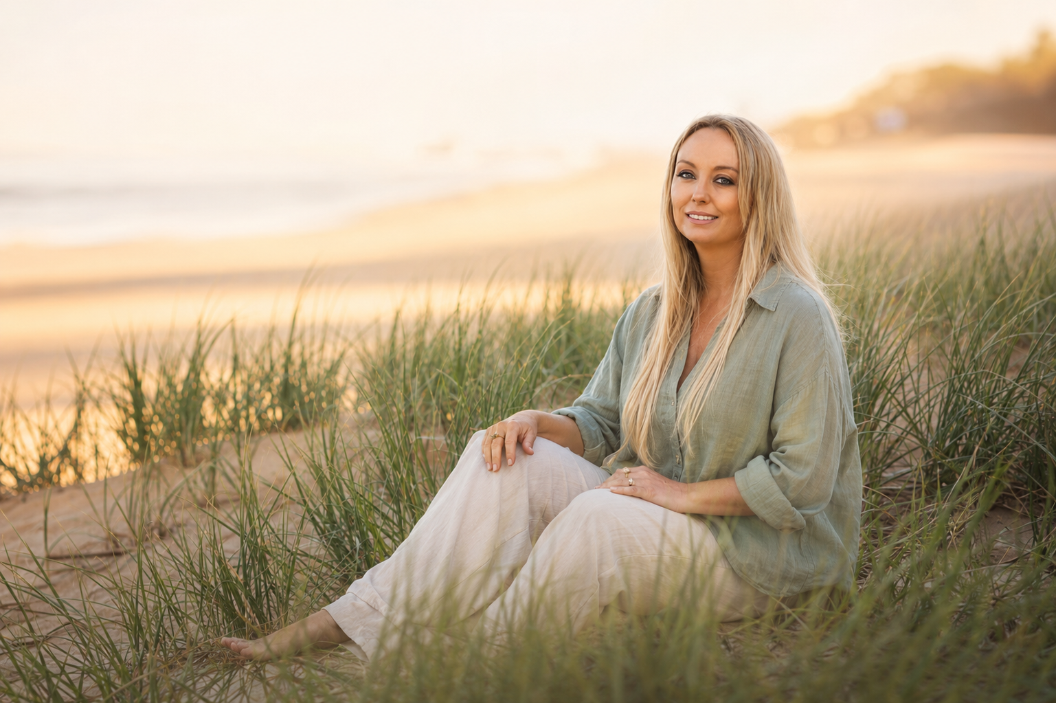 Blonde woman sitting in beach grass at sunset, wearing a sage-green shirt and white pants.