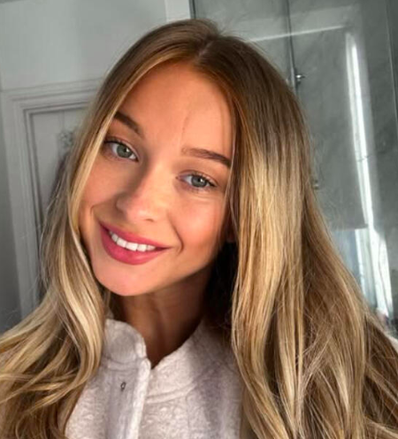 Close-up of a young woman with long blonde hair and blue eyes, smiling indoors.