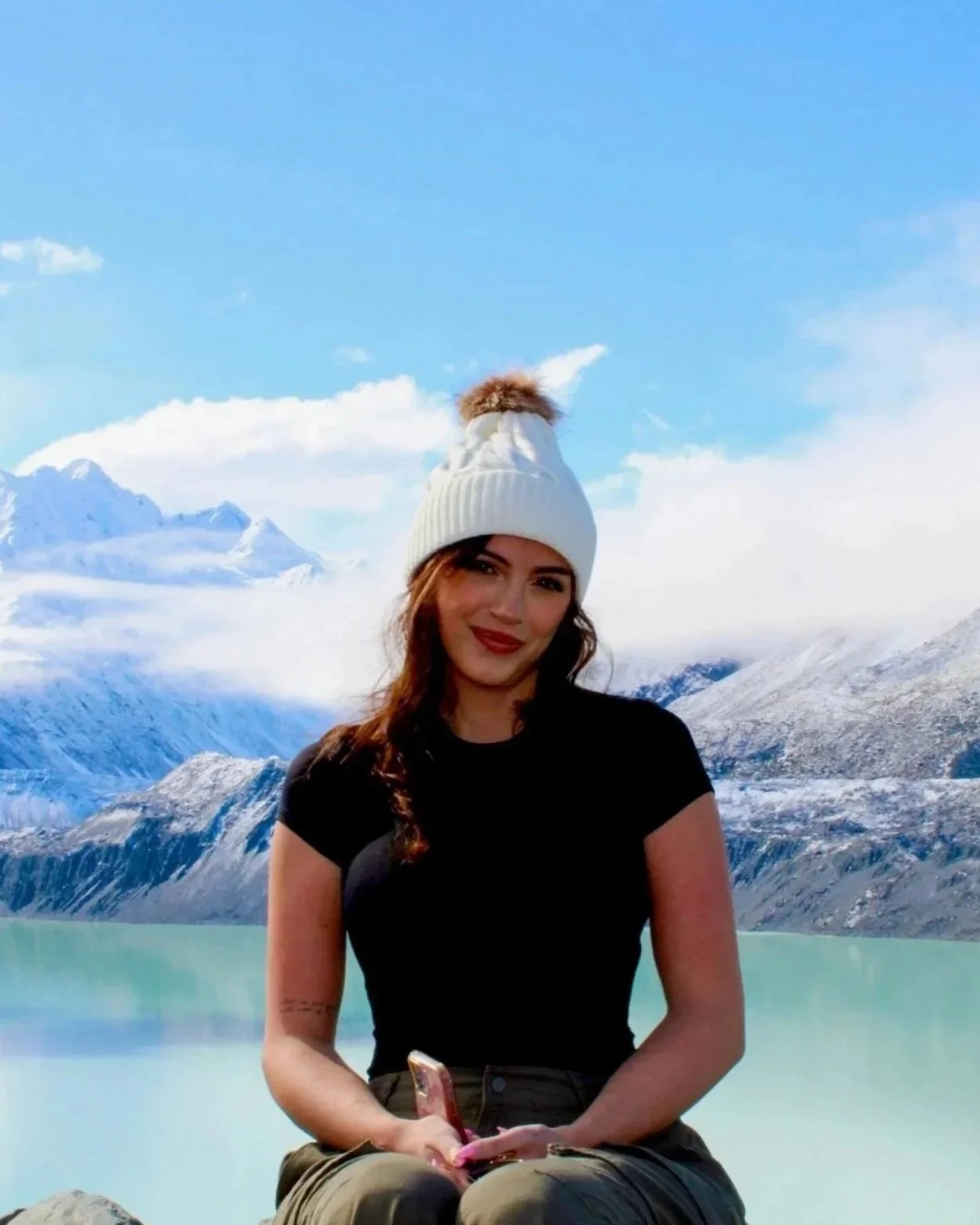 Woman in black shirt and white knit hat with pom-pom sitting outdoors near a lake with mountains and snow behind her.