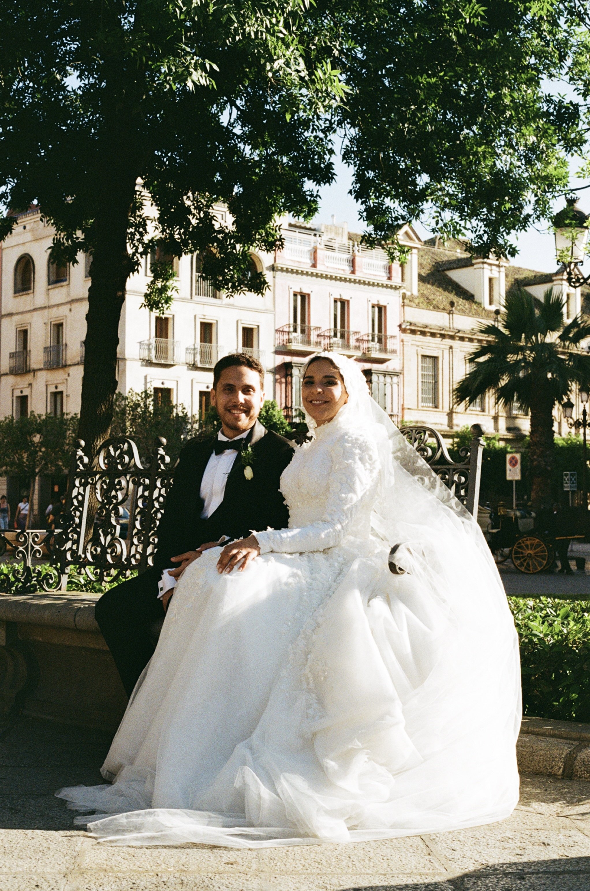 A bride and groom sitting on a park bench, smiling for the camera. The bride is wearing a white wedding gown with a long veil, and the groom is wearing a black tuxedo with a bow tie. There are trees and buildings in the background, suggesting an outd
