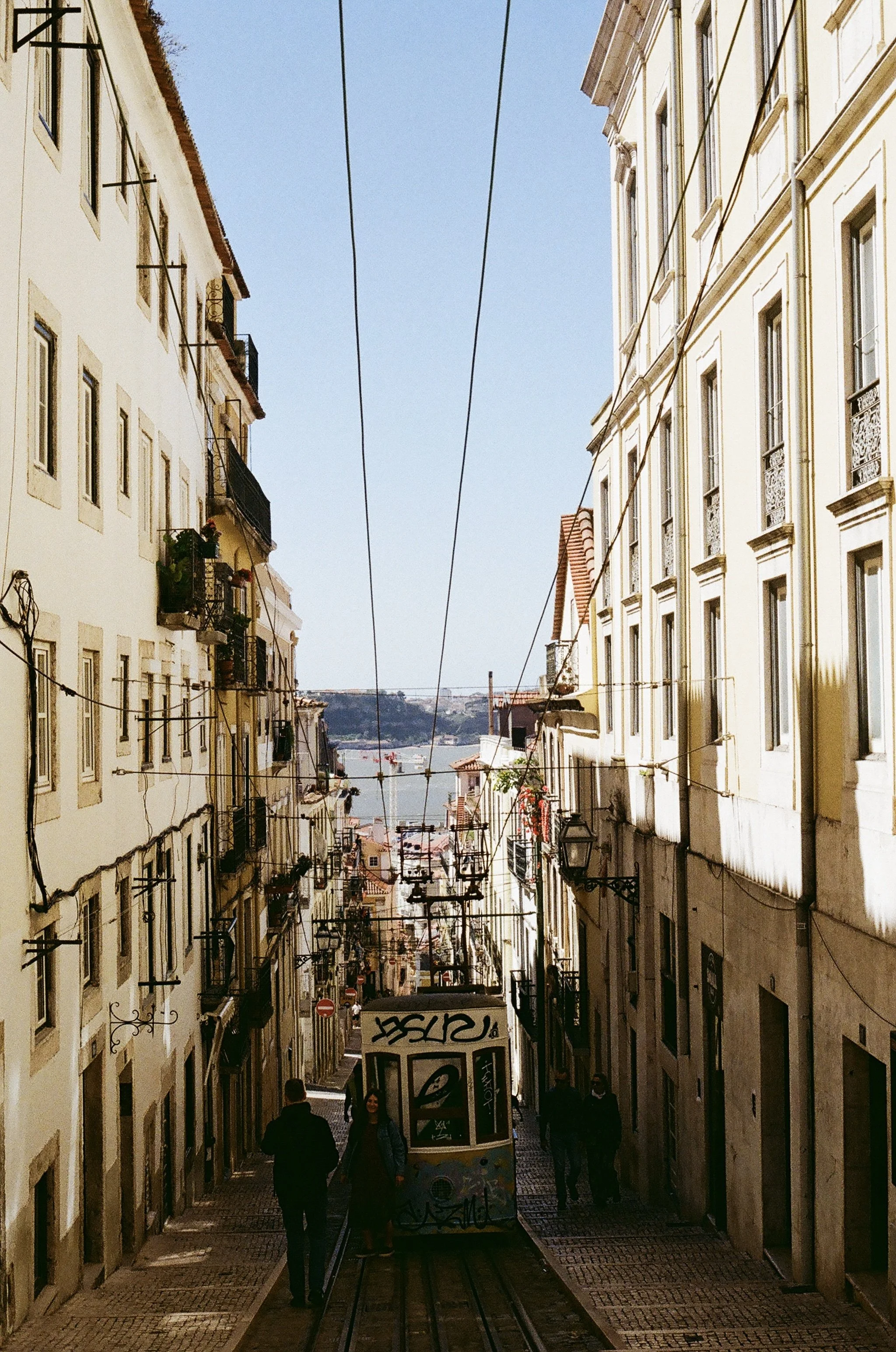 City street with tram, people walking, tall buildings on both sides, and a view of water and ships in the distance. The street is narrow and lined with streetlights and overhead wires for the tram.