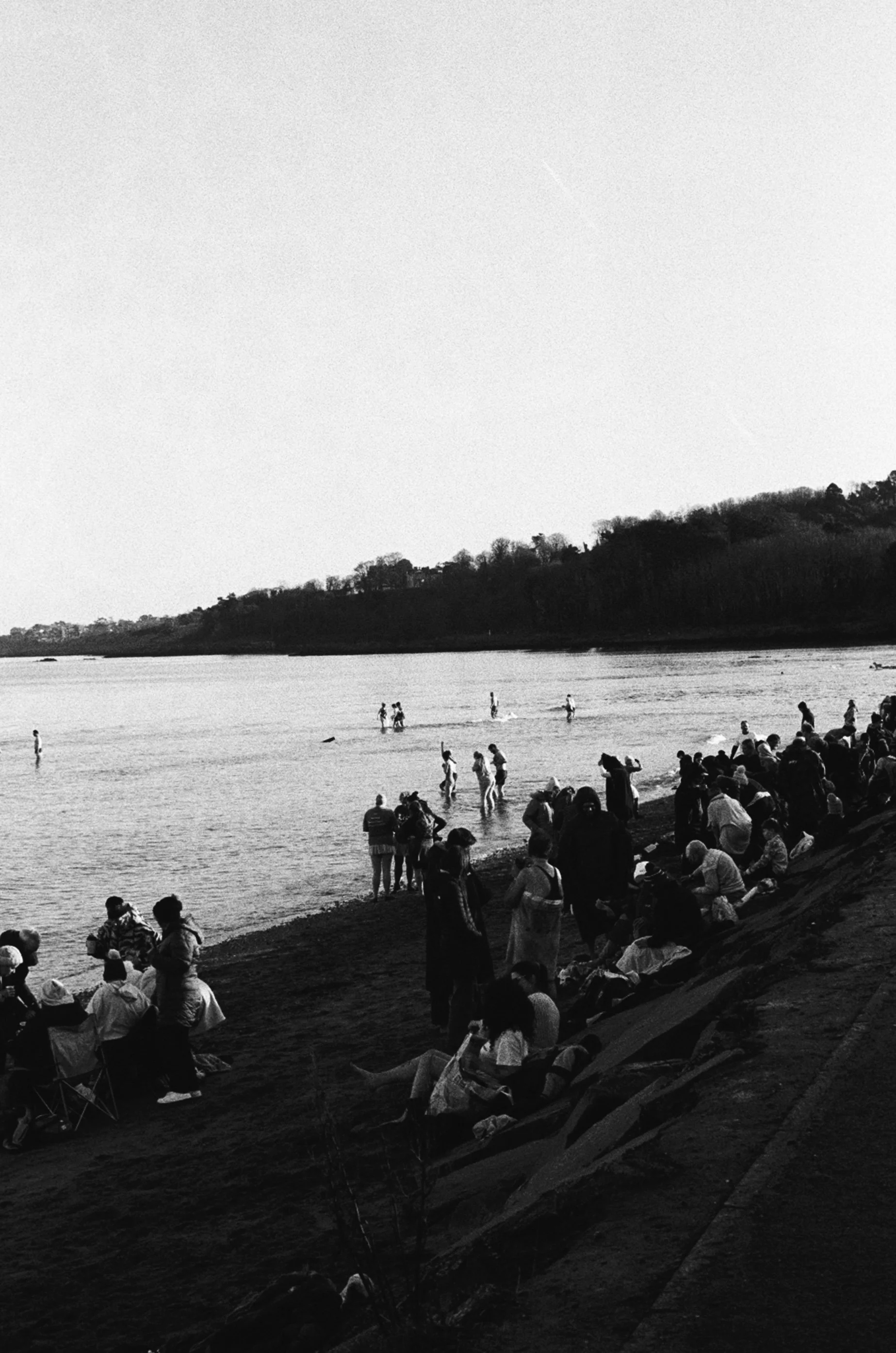 Black and white photo of a crowded beach with people relaxing on the sand and swimming in the water, and hills in the background.