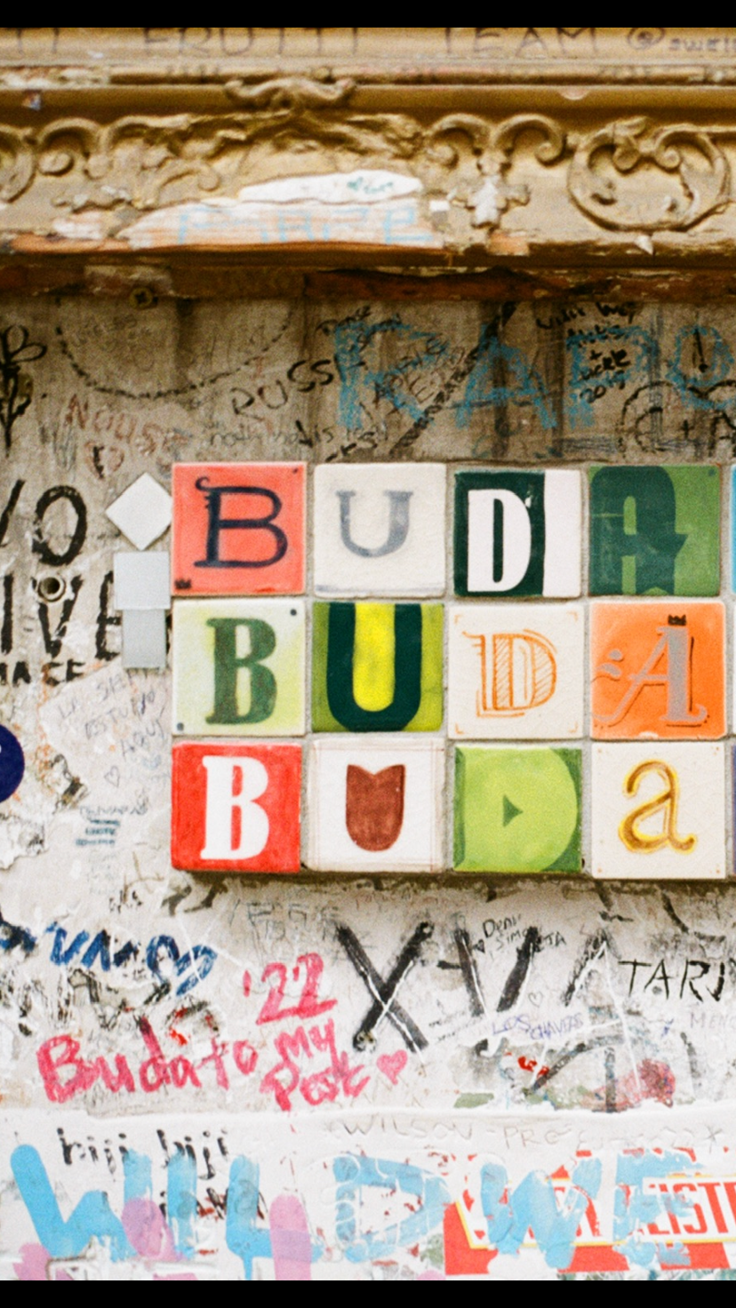 Colorful tiles spelling 'BUDABU', attached to a graffiti-covered wall.