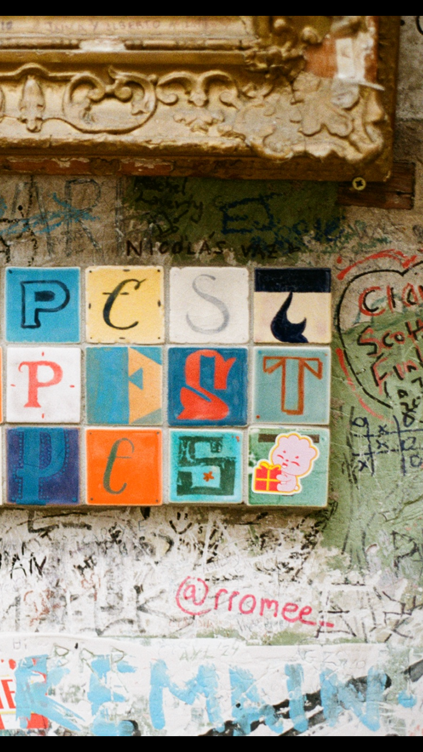 Colorful tiles with letters P, C, S, T, and images including a pink character holding a gift, arranged on a graffiti-covered wall.