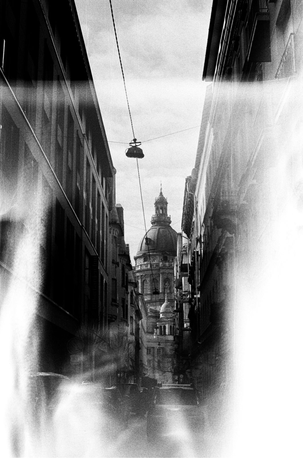 Black and white photo of a narrow city street with a domed building in the background, overhead wires, and some blurred elements and light streaks.
