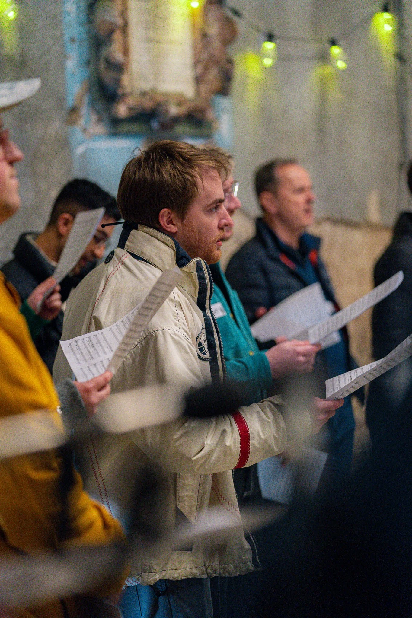 Group of people holding sheet music or scripts, standing indoors with string lights, a rustic wall, and a decorative figure in the background.