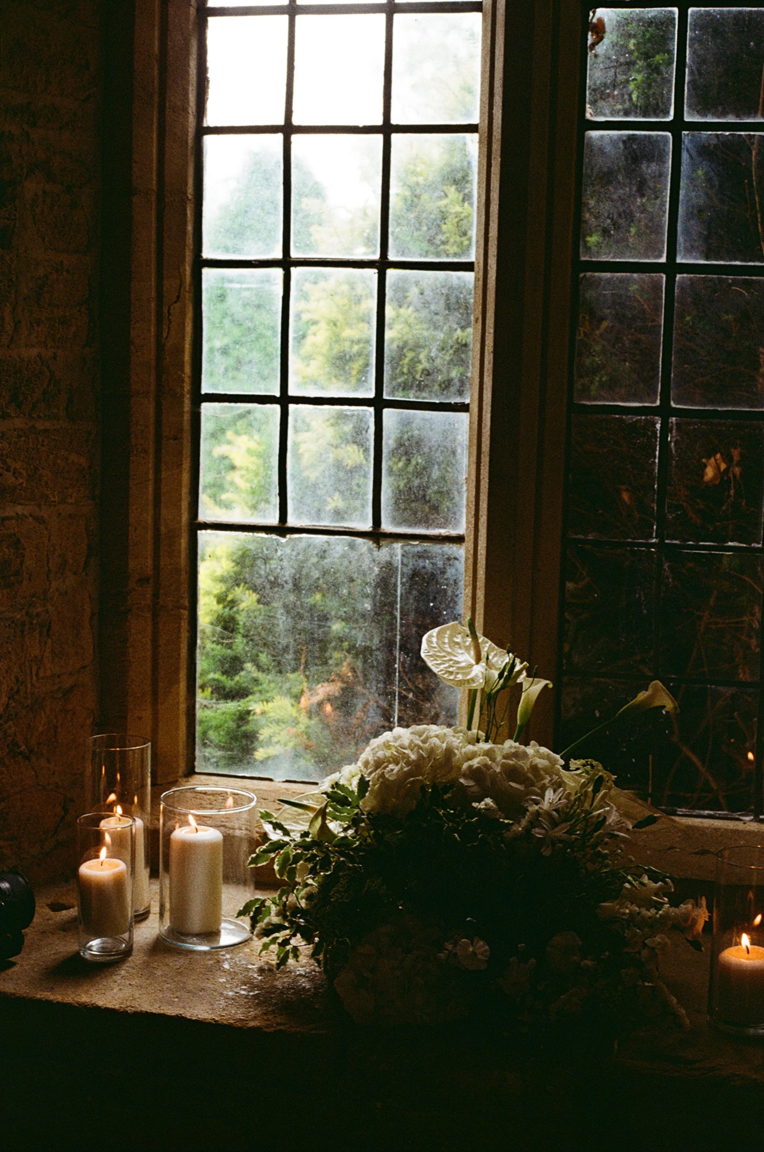 A window with an outdoor view of trees, with a windowsill decorated with candles and white flowers.
