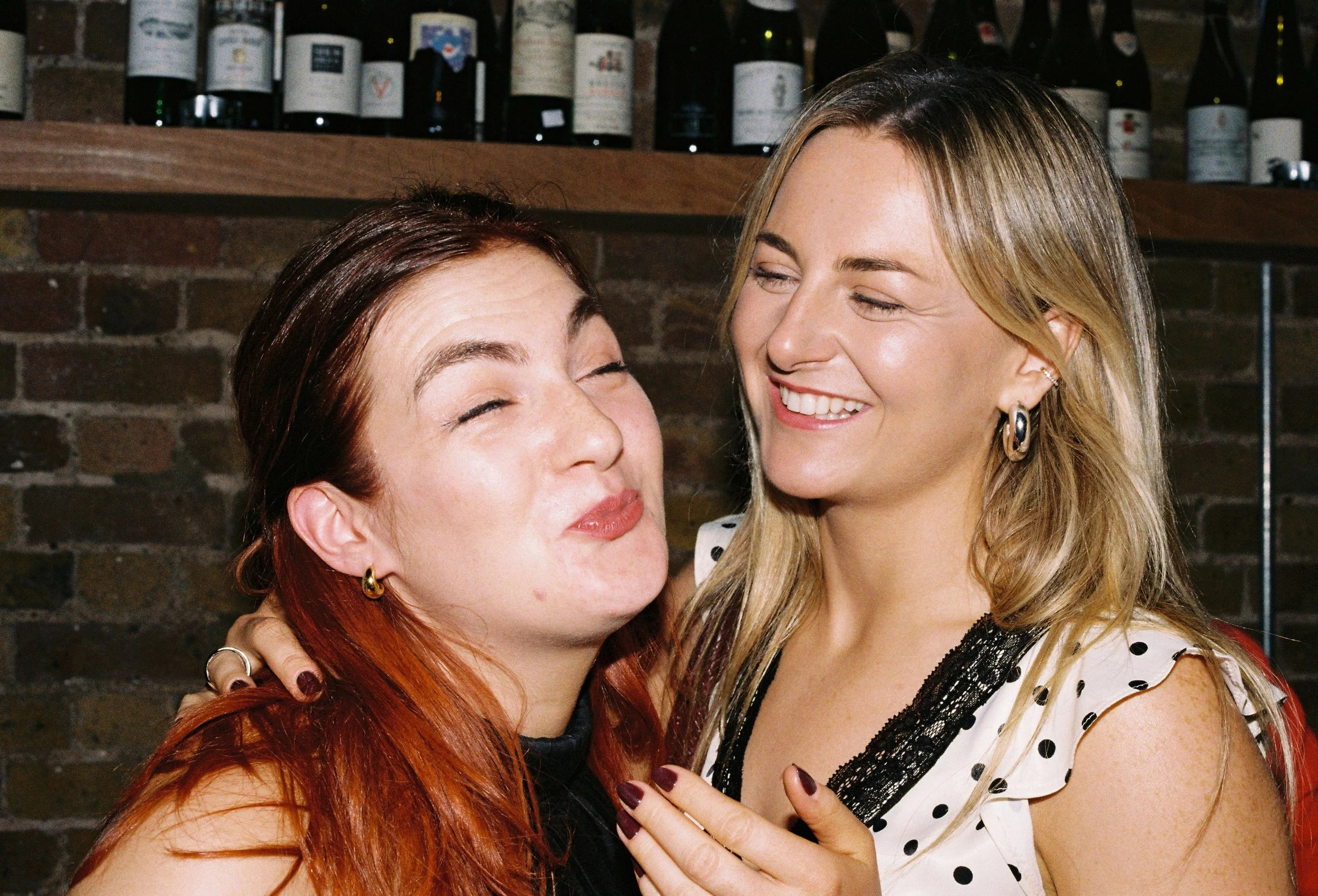 Two women smiling and embracing each other in a friendly and joyful moment in a restaurant or bar.