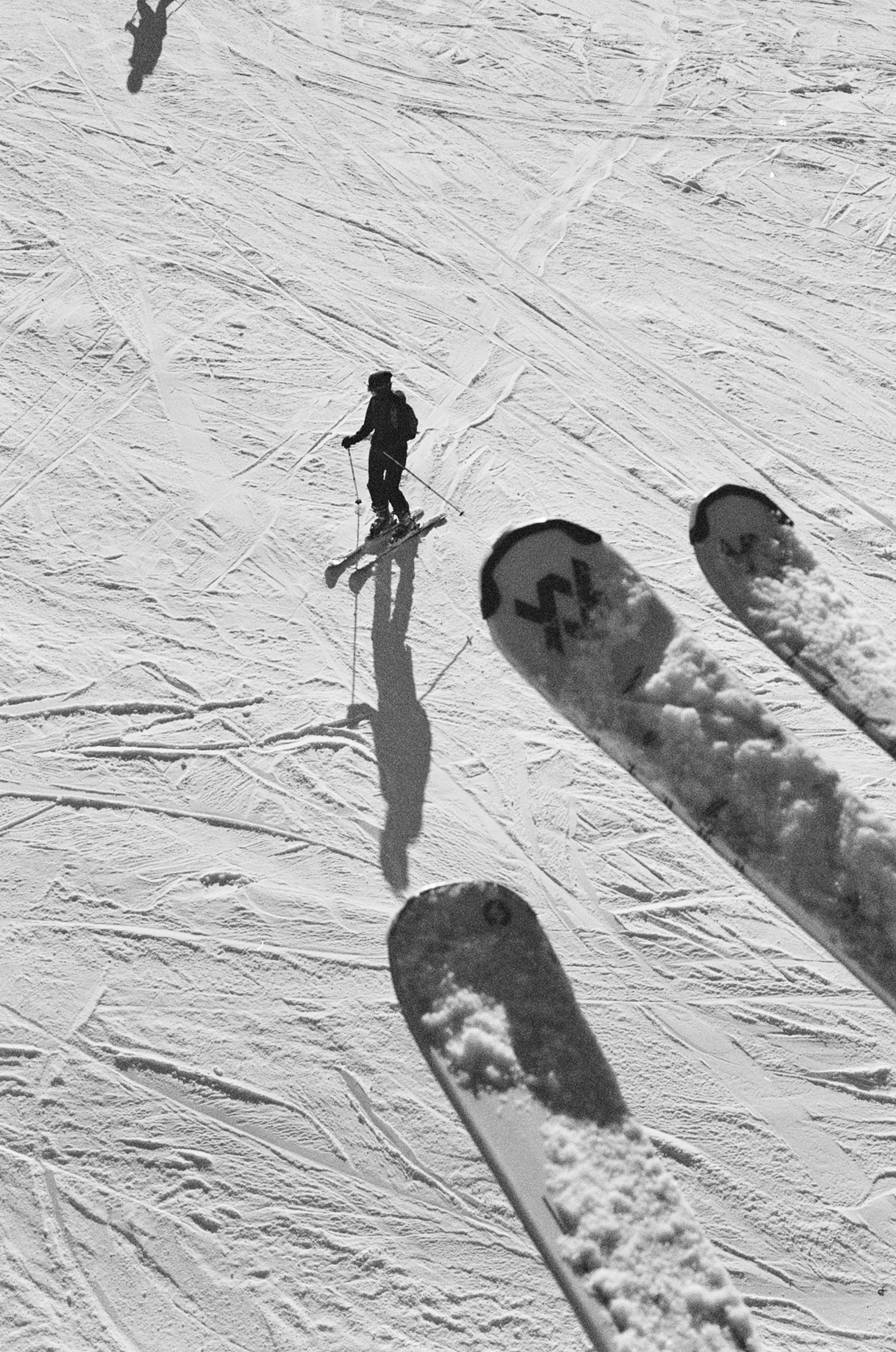 A skier in a black outfit skiing downhill on snow with ski tracks around, viewed from above, with the shadow of the skier visible on the snow, and two skis with snow on top in the foreground.