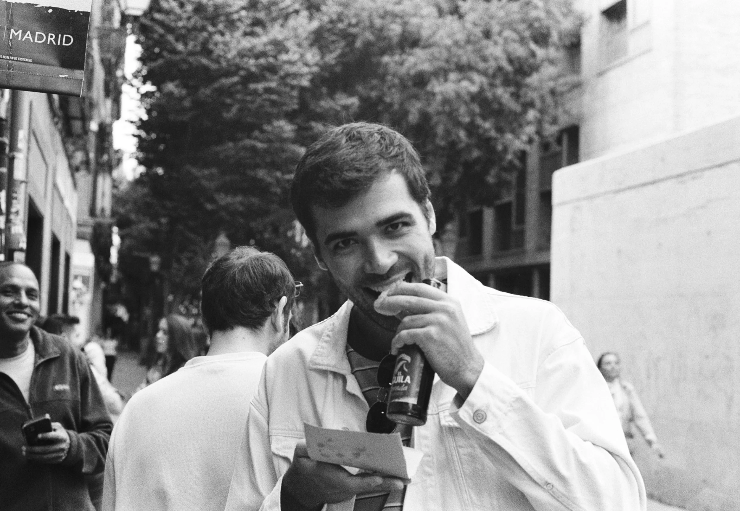 A group of people standing on a city street, with one young man in the foreground smiling and drinking from a bottle, holding a piece of paper. Others in the background are talking and smiling.