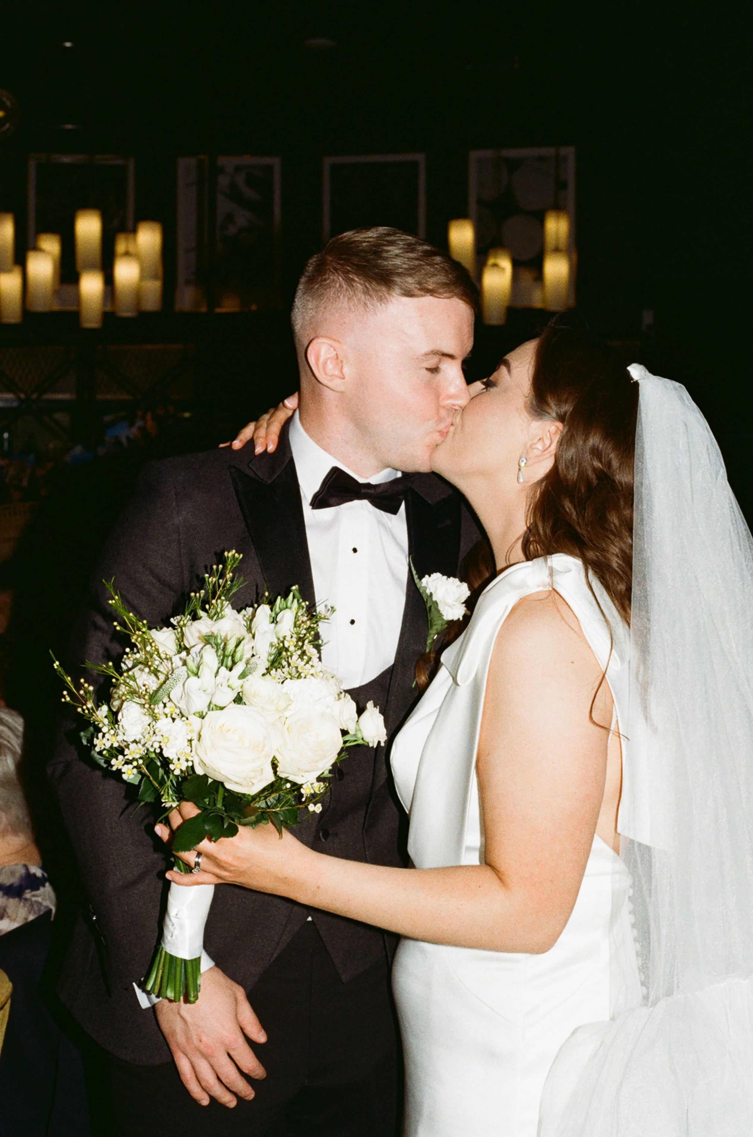 A bride and groom share a kiss at their wedding, with the groom in a tuxedo holding a bouquet of white flowers.