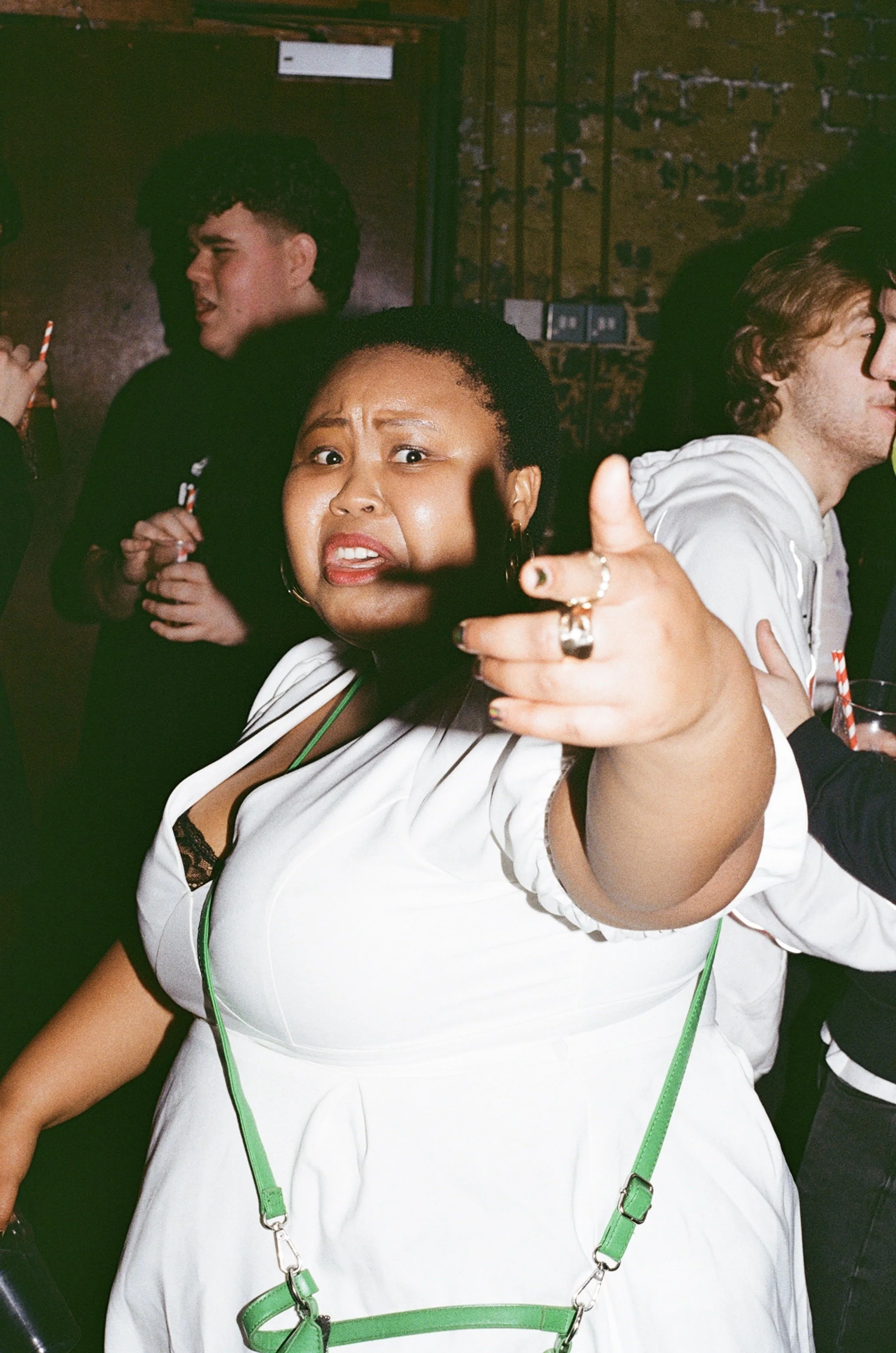 A woman in a white outfit with a green purse points towards the camera with a surprised or confused expression, surrounded by people at a social gathering.