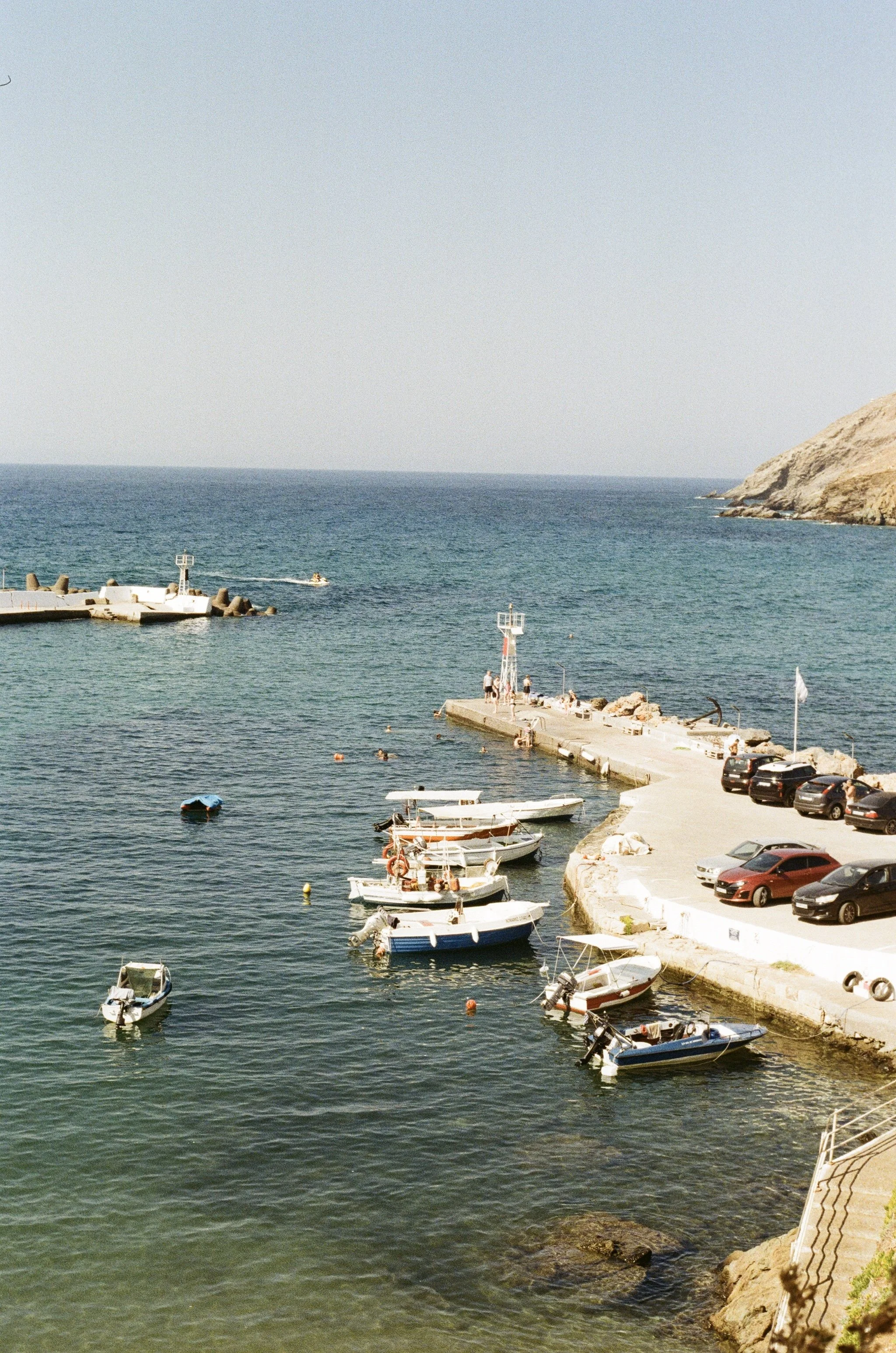 A small harbor with boats docked in the water, a concrete pier extending into the sea, parking area with cars, rocky coastline, and a clear blue sky.