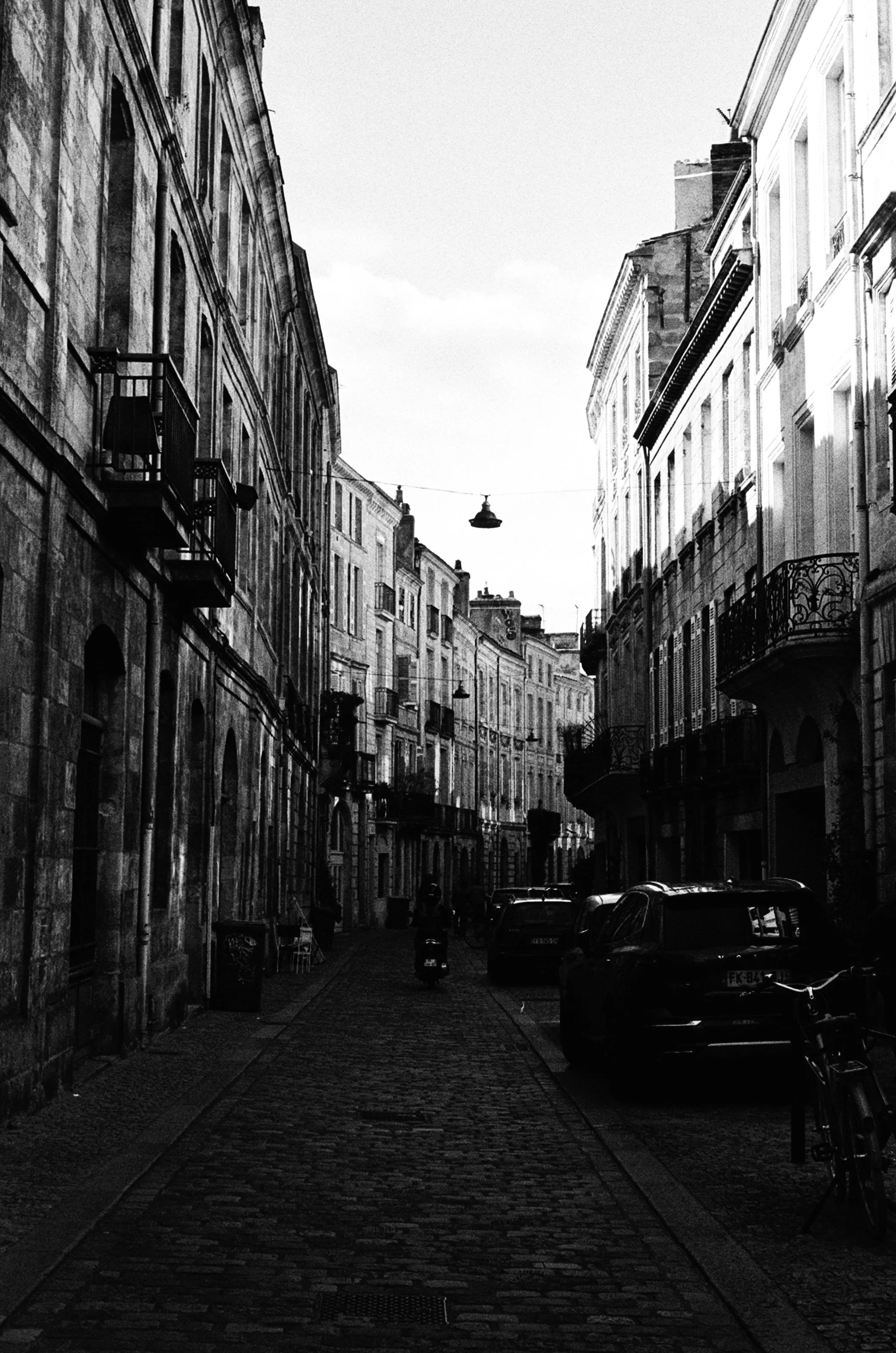Black and white photo of a narrow European street with cobblestone pavement, tall historic buildings on both sides with balconies and ornate details, parked cars, bicycles, and a person riding a scooter.