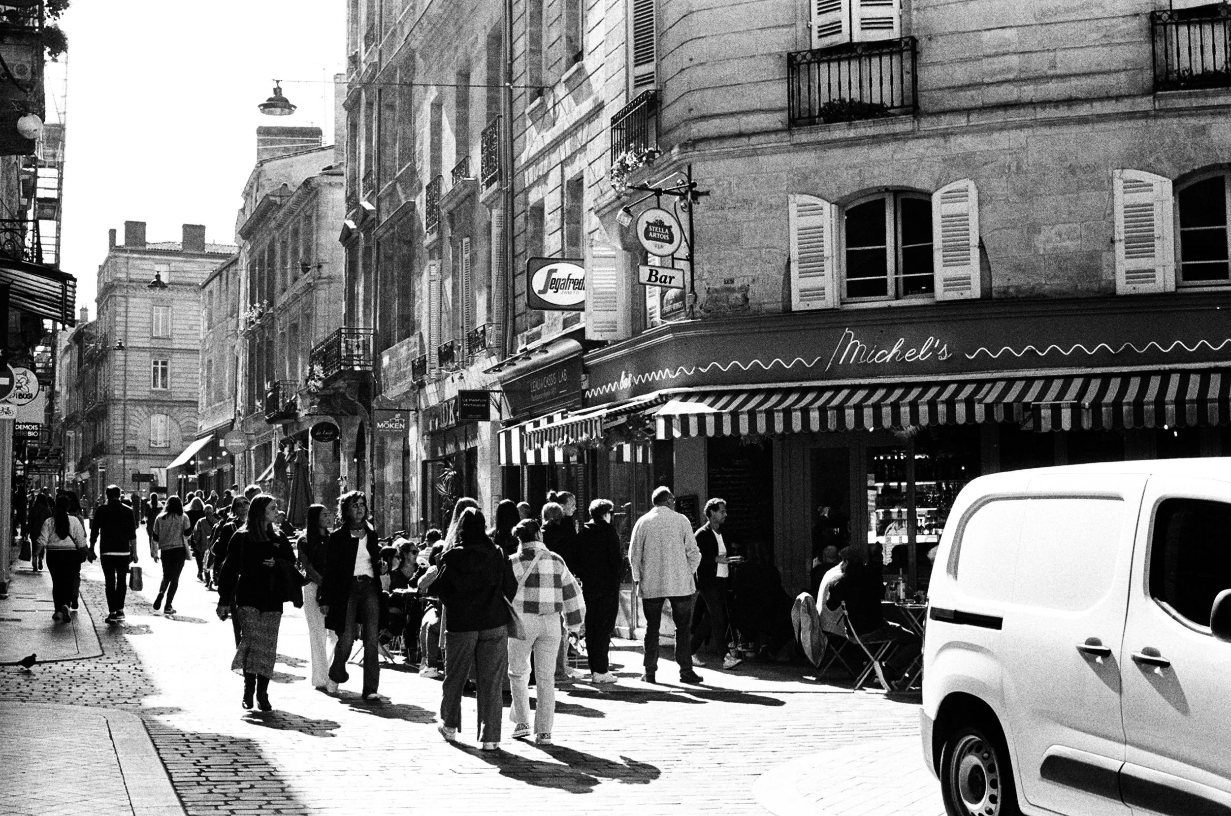 Black and white photo of a busy European city street with people walking on the sidewalk, some sitting outside a cafe named Michel's, and a white van in the foreground.