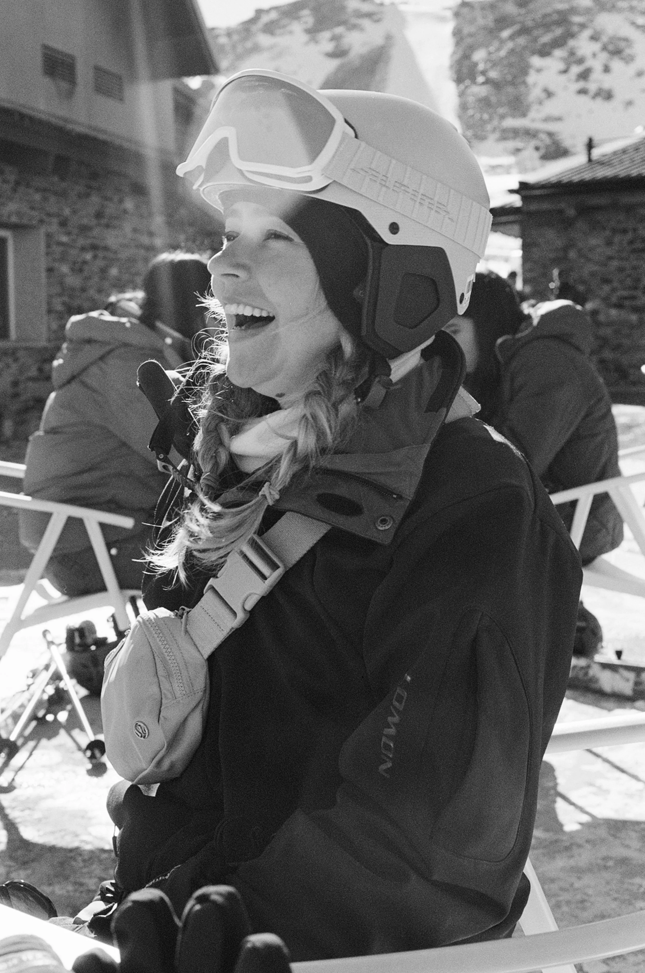 A woman wearing a ski helmet and goggles, smiling while looking to the side, with a mountain and ski resort buildings in the background.