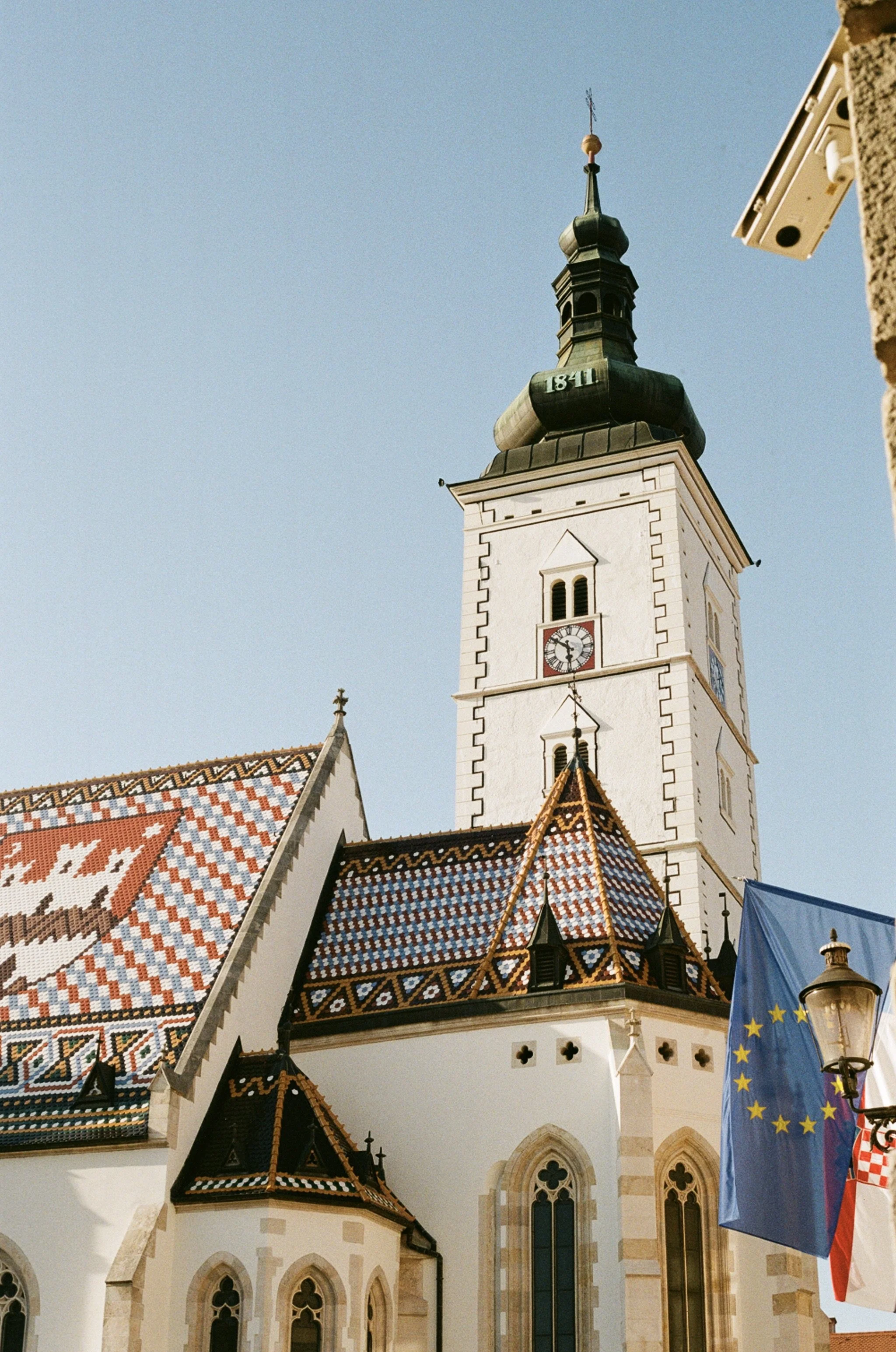 A church with a tall clock tower, a colorful tiled roof, and a European Union flag and Croatian flag in front.