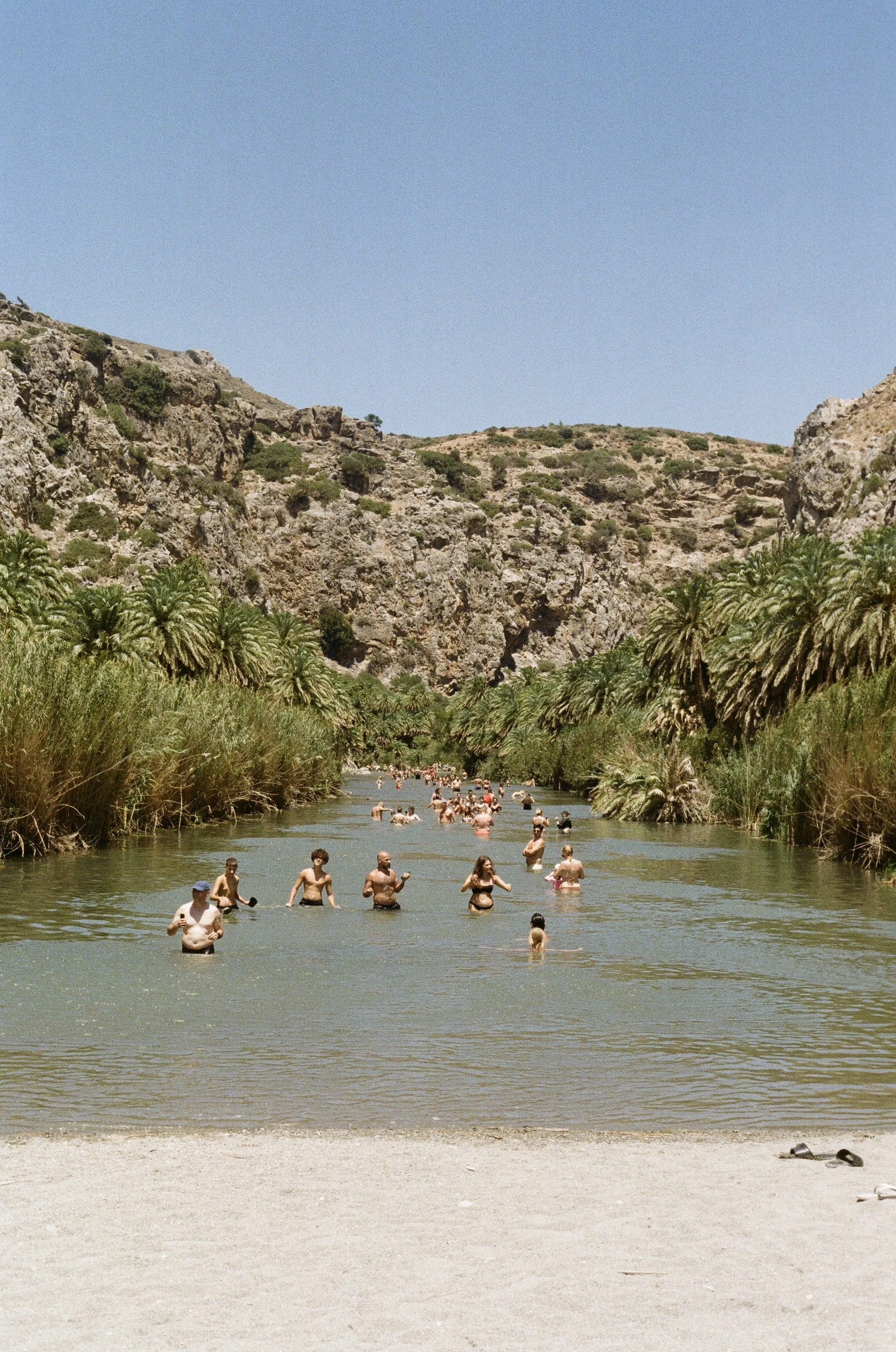 People swimming and relaxing in a river on a sunny day, surrounded by palm trees and rocky hills.