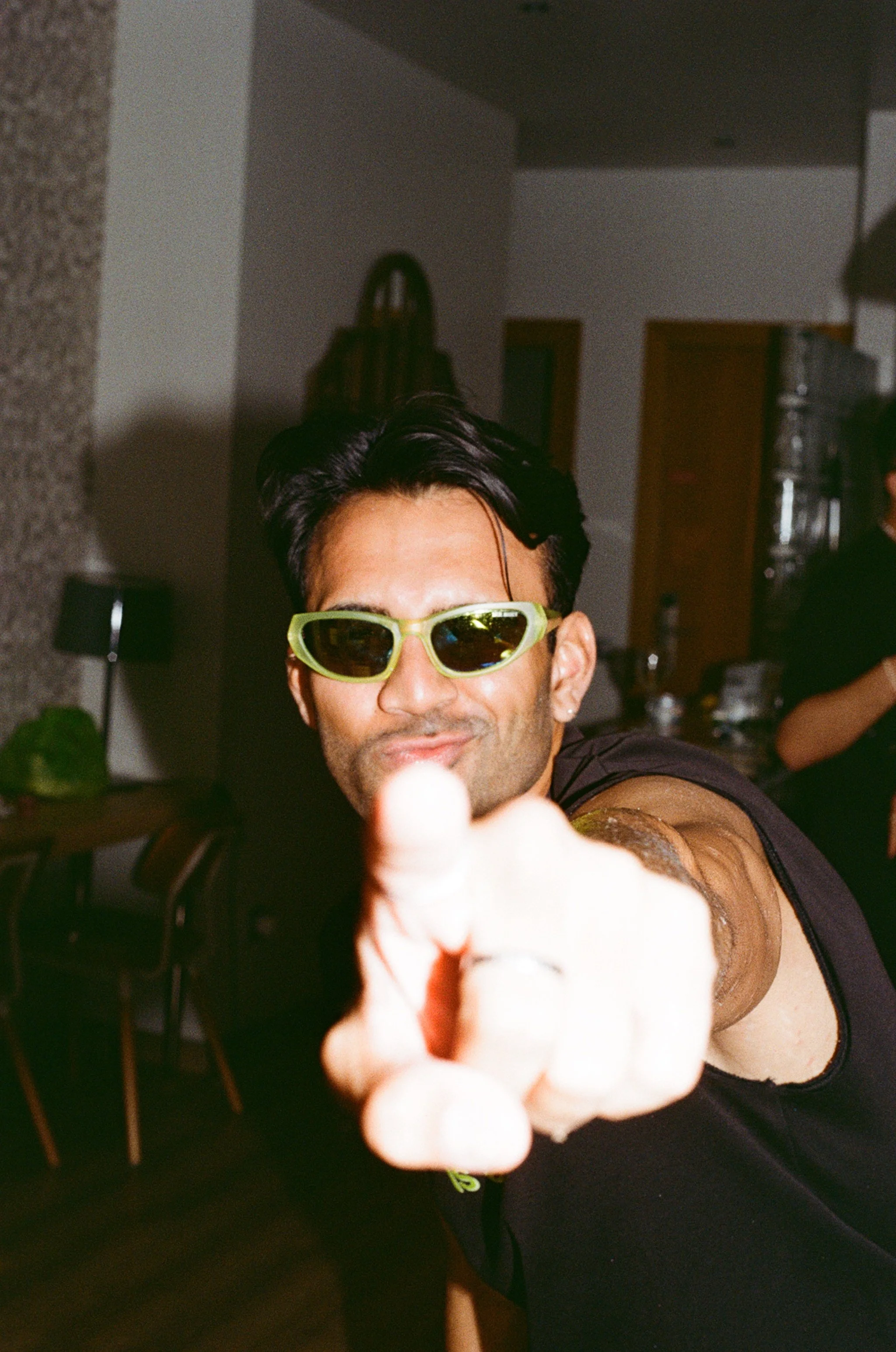 A man wearing yellow sunglasses, smiling, and reaching toward the camera in an indoor setting.