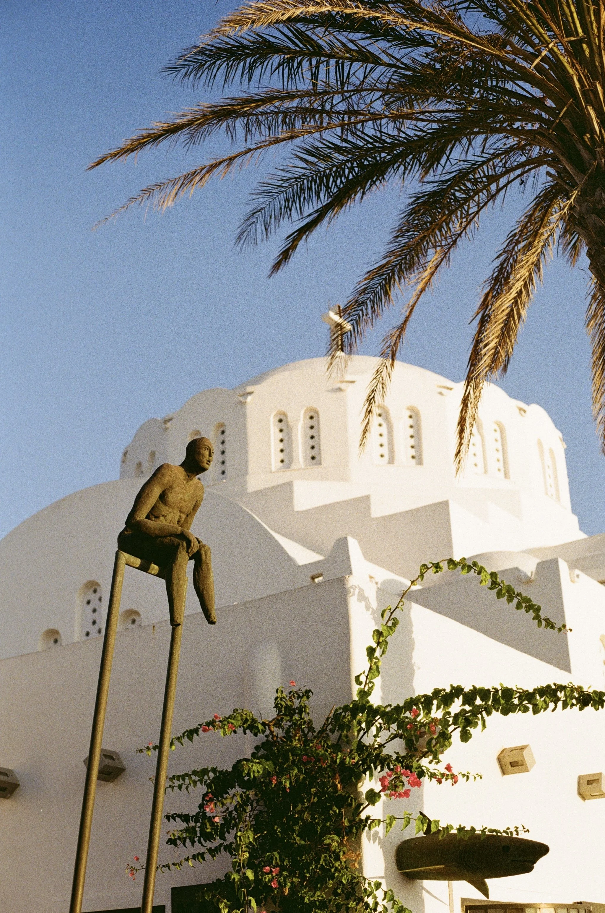 A white domed building with small arched windows, a palm tree with long fronds in the foreground, a small sculpture of a seated figure on tall stilts, and flowering plants.