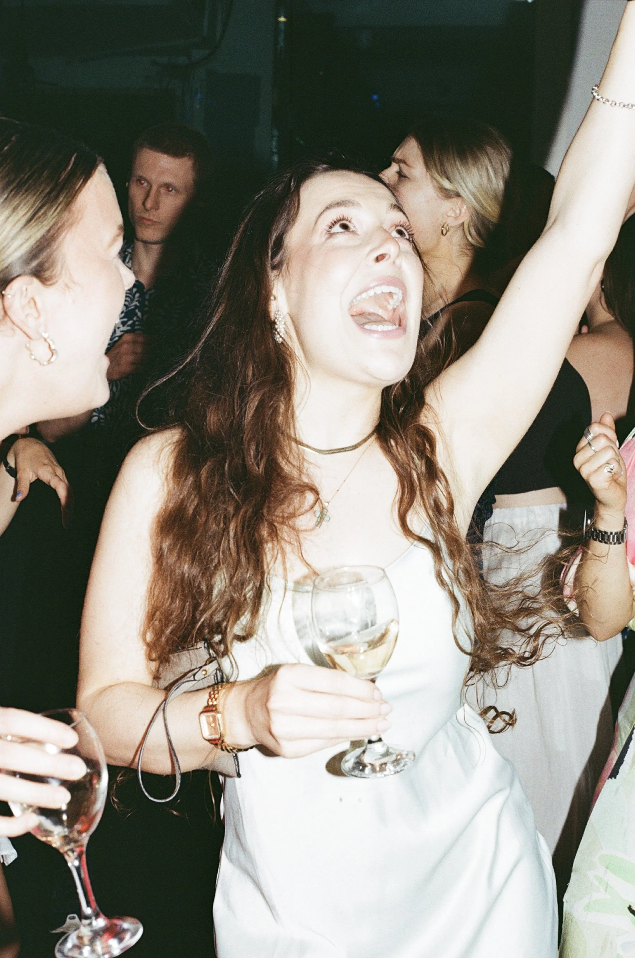 A woman with long, curly red hair holding a glass of white wine at a celebration or party, surrounded by other people who are also enjoying the event.