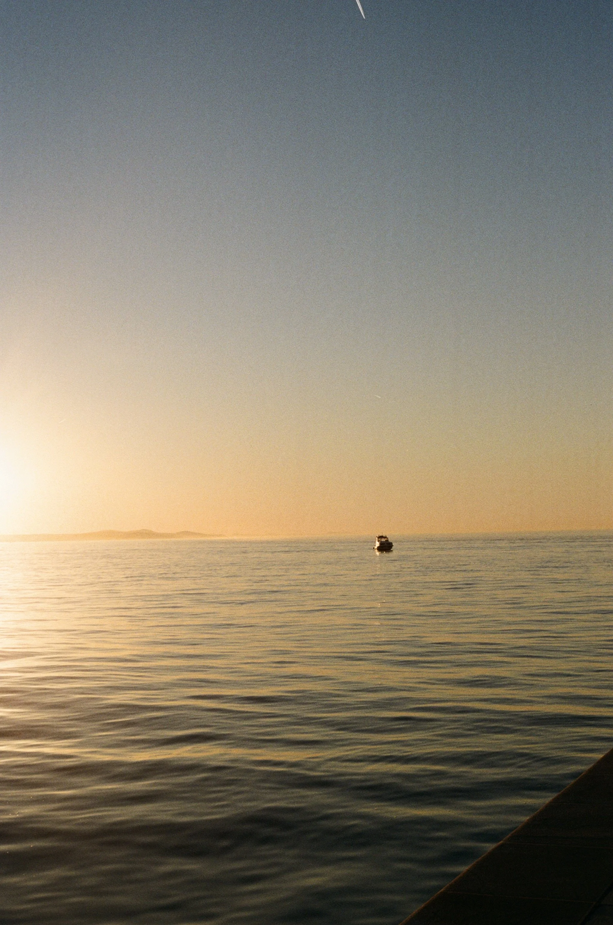 Sunset over calm ocean with a small boat in the distance, partially visible shoreline on the right.