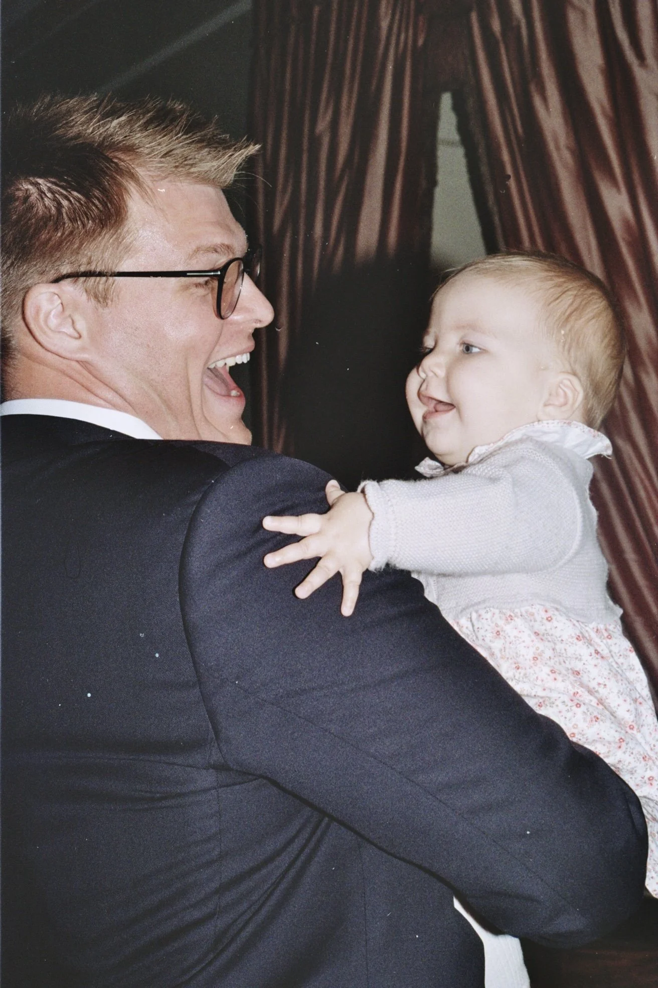 A man in a suit and glasses holding a baby girl, both smiling at each other.