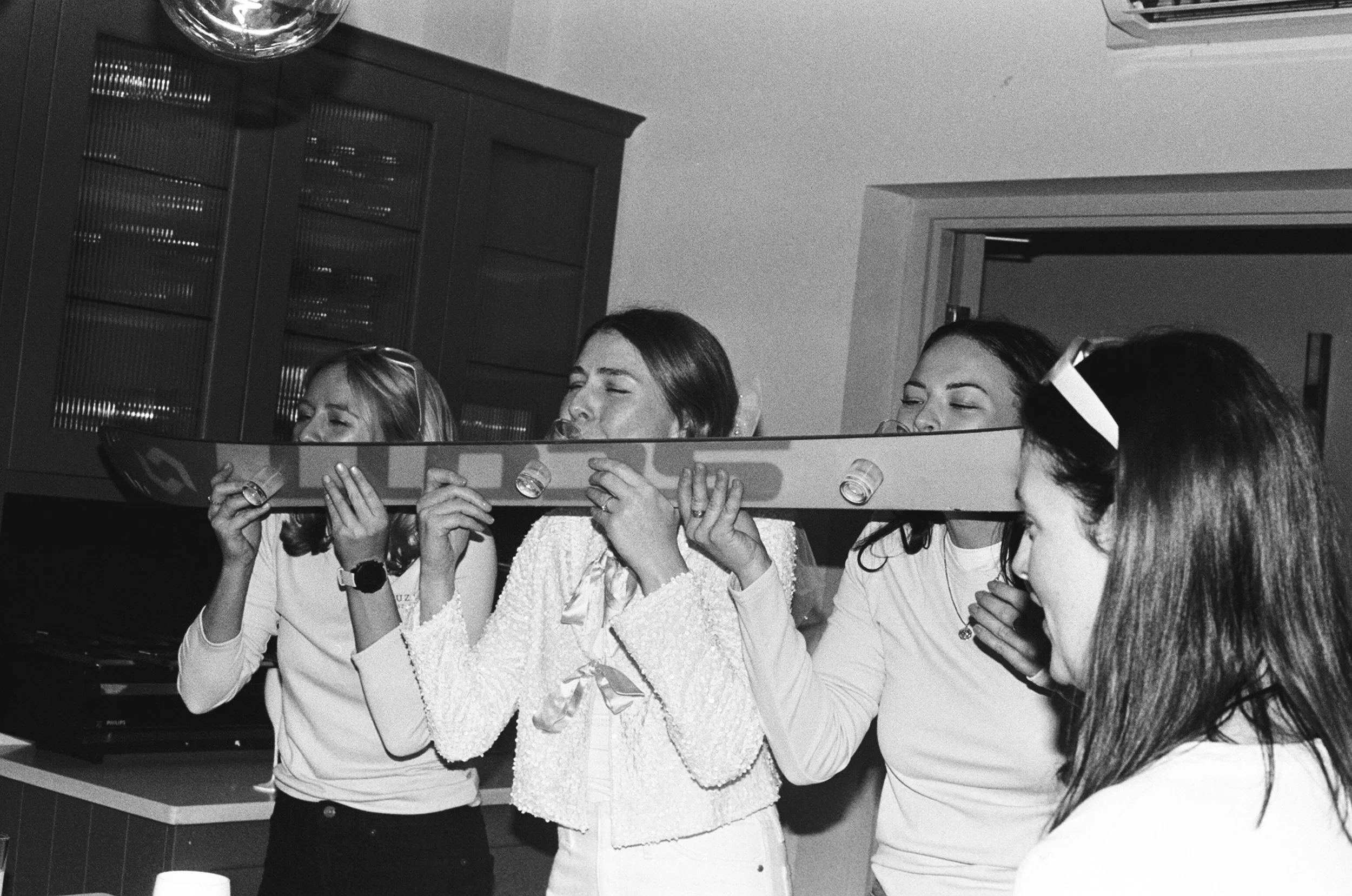 Four women celebrating with a large elongated toast, with no visible glasses or drinks, in a kitchen or dining area.