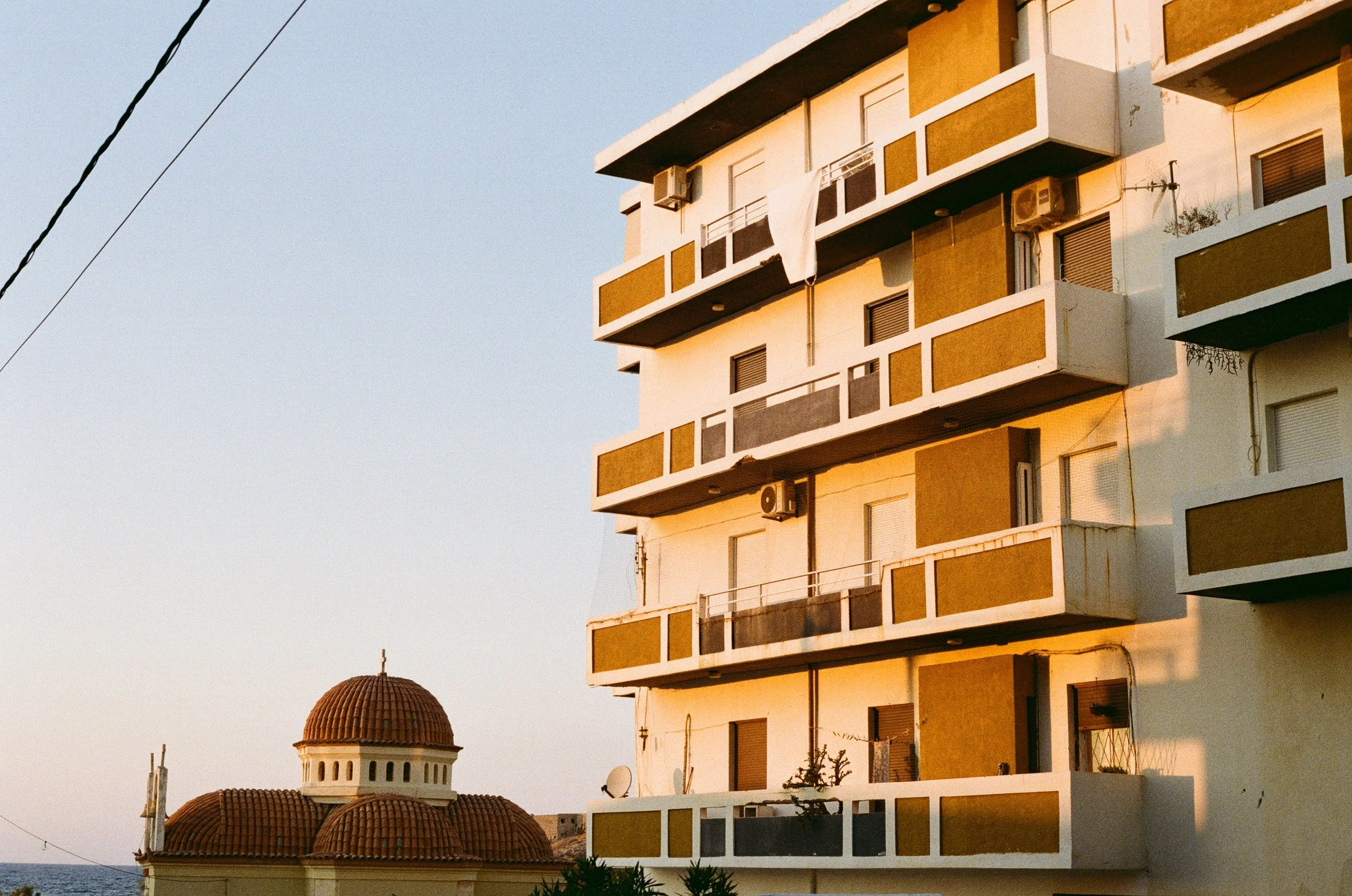 A multi-story apartment building with balconies, some with air conditioning units, and a white cloth hanging on one of the balconies. In the background, there is a building with a red-tiled dome roof, likely a church or similar structure. The scene i