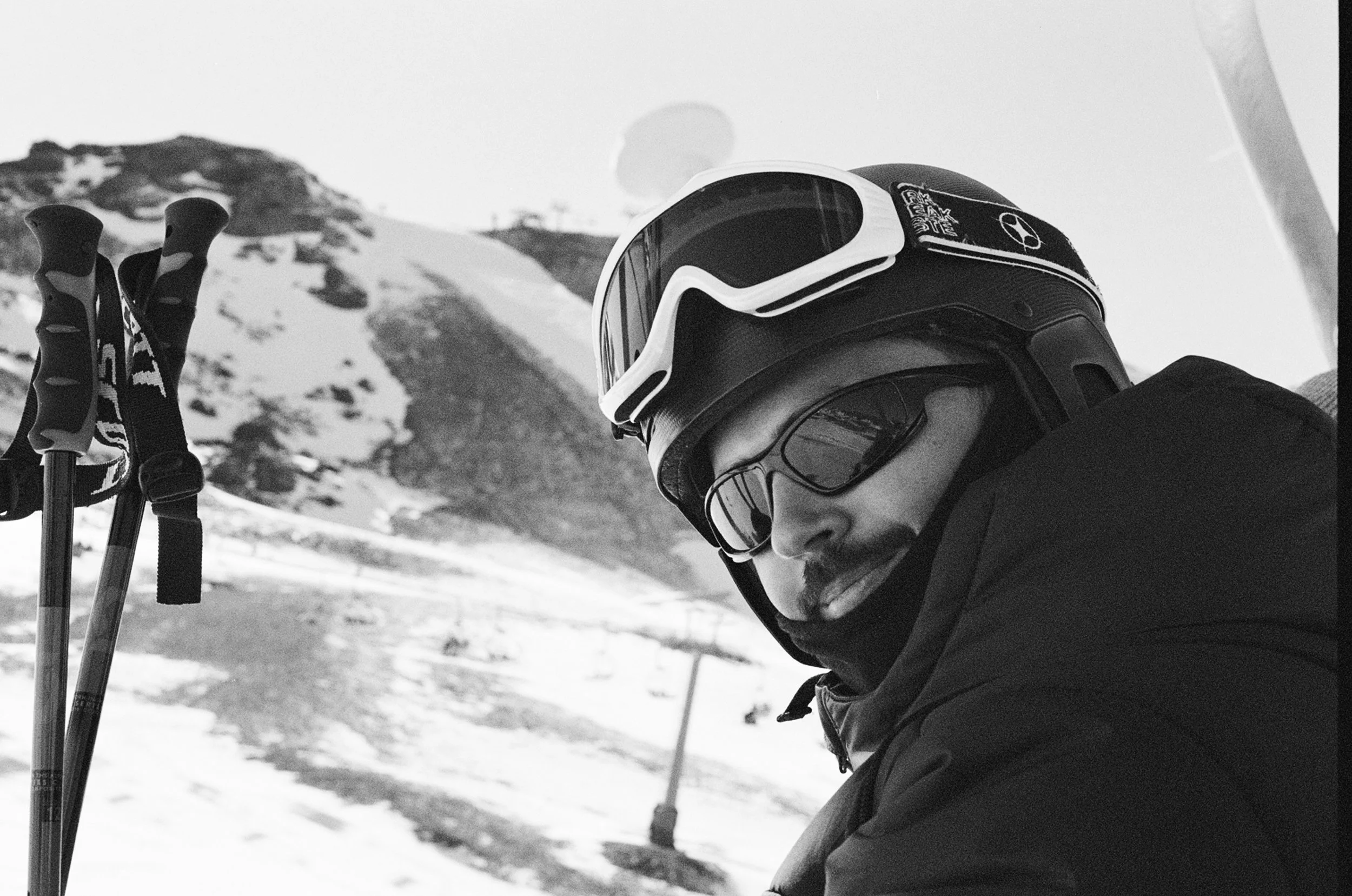 A man wearing a ski helmet, goggles, and winter gear sits with snow-covered mountain in the background.