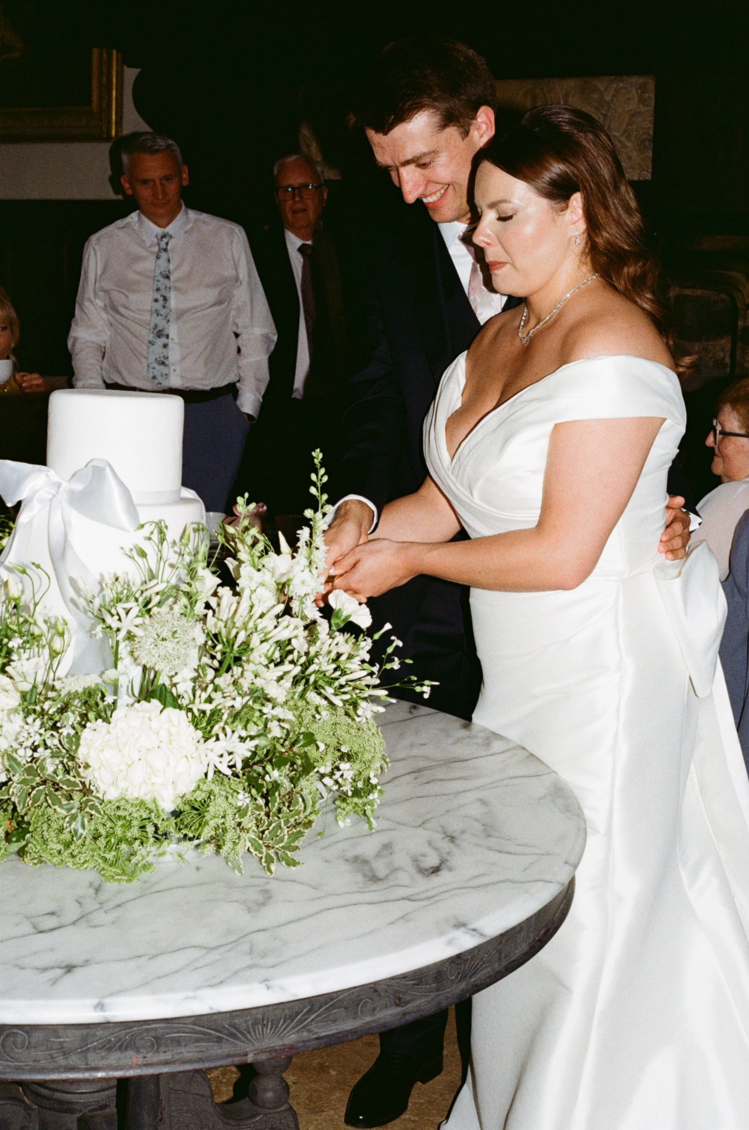 Bride and groom cutting wedding cake at reception, surrounded by guests, with a floral arrangement on a marble table.