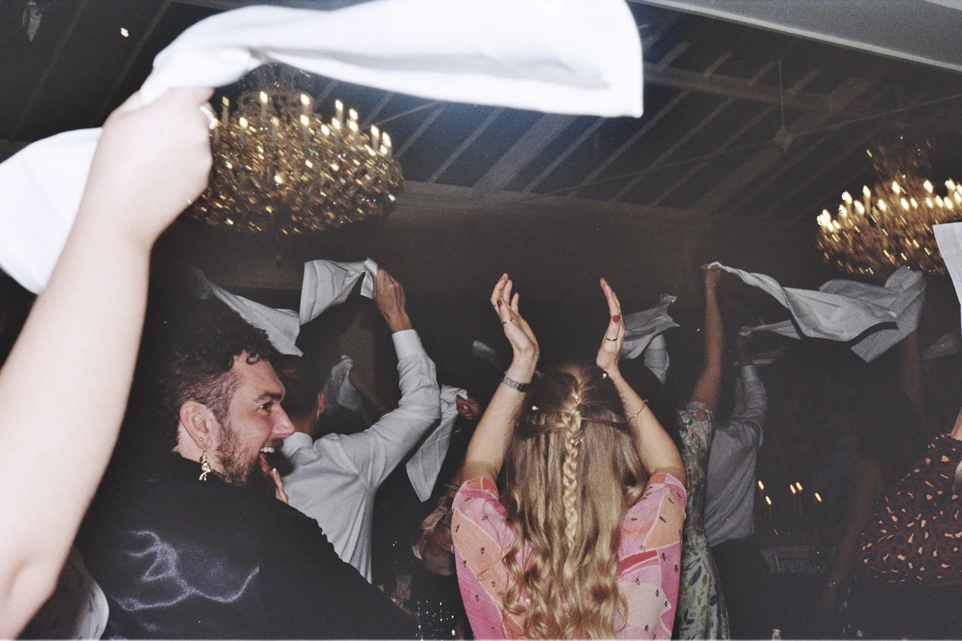 People dancing and celebrating at a party in a dimly lit room with chandeliers.