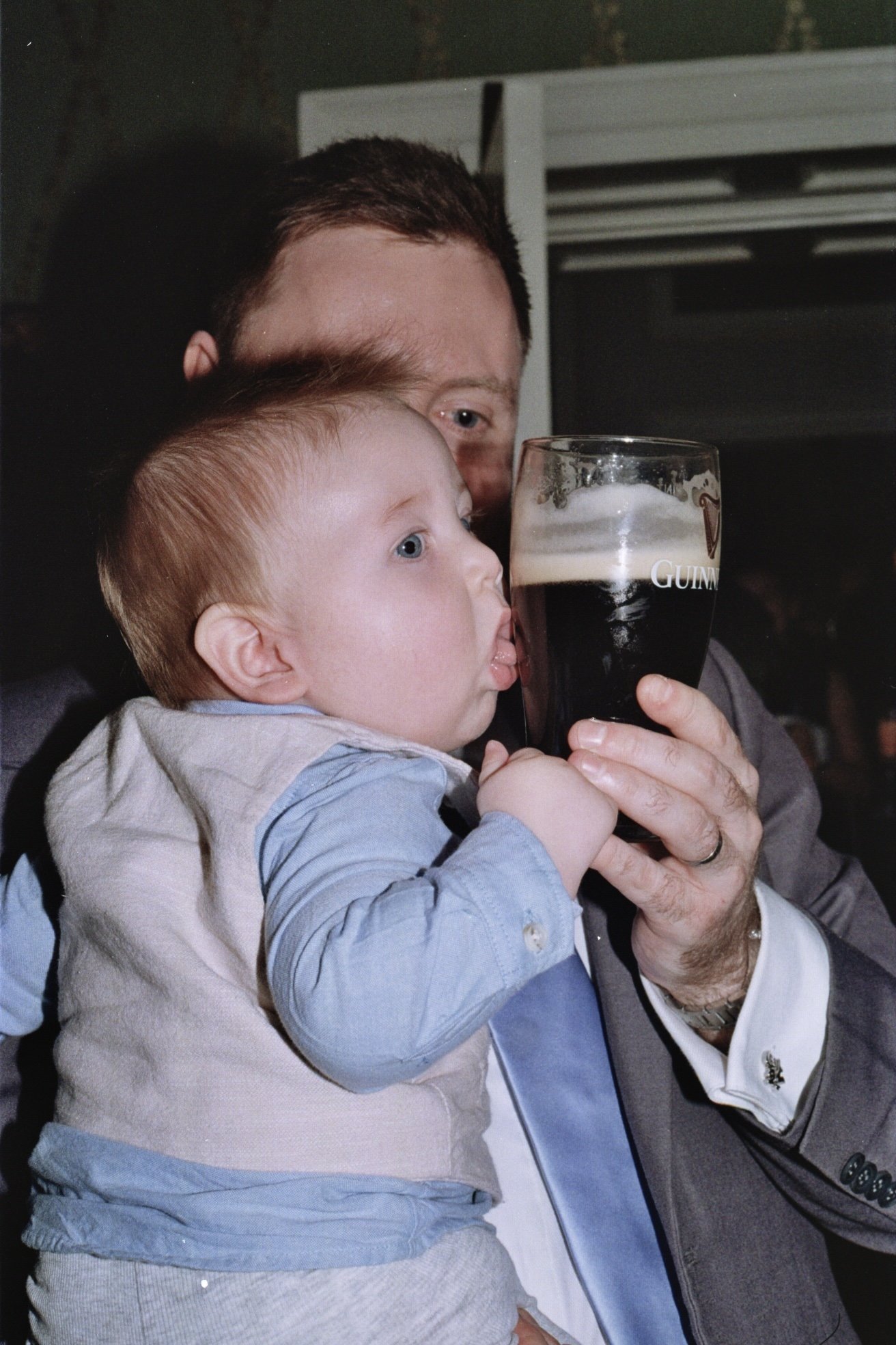 A man holding a young child in front of his face, both tasting a pint of dark stout beer in a pub.