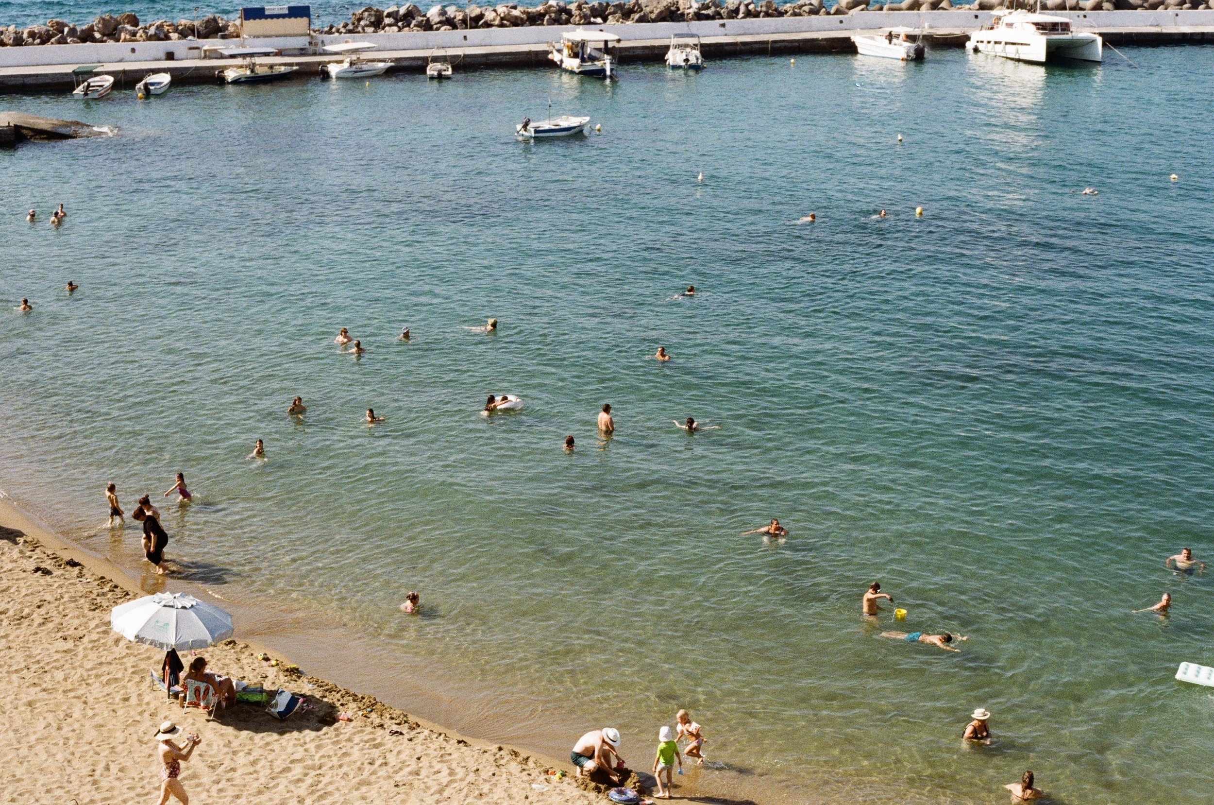 Beach scene with people swimming in the ocean and relaxing on the sandy shore, some under umbrellas, near a marina with boats docked.
