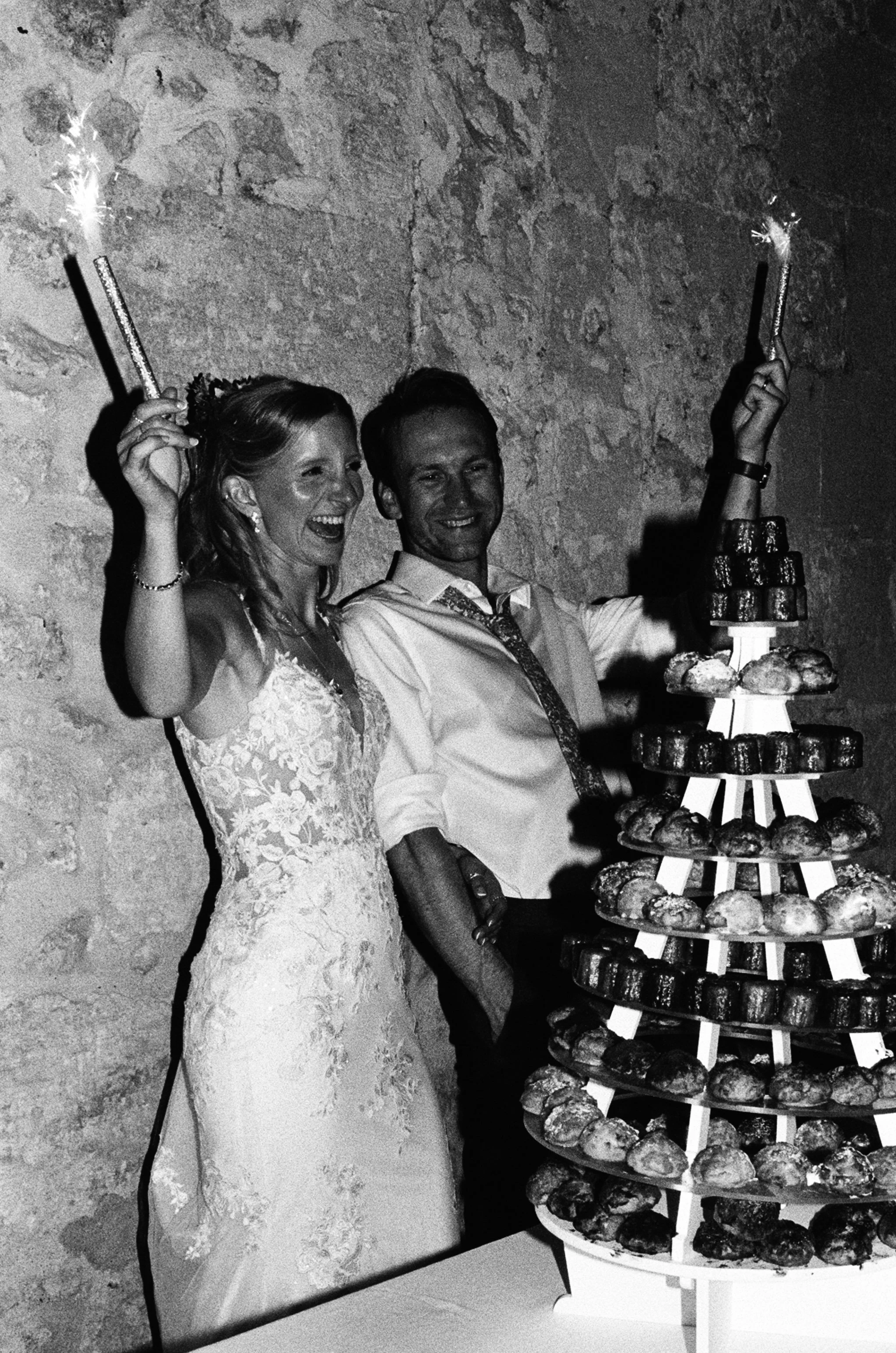 A newlywed couple celebrating their wedding with sparklers, standing next to a tiered display of pastries or donuts at a wedding reception.