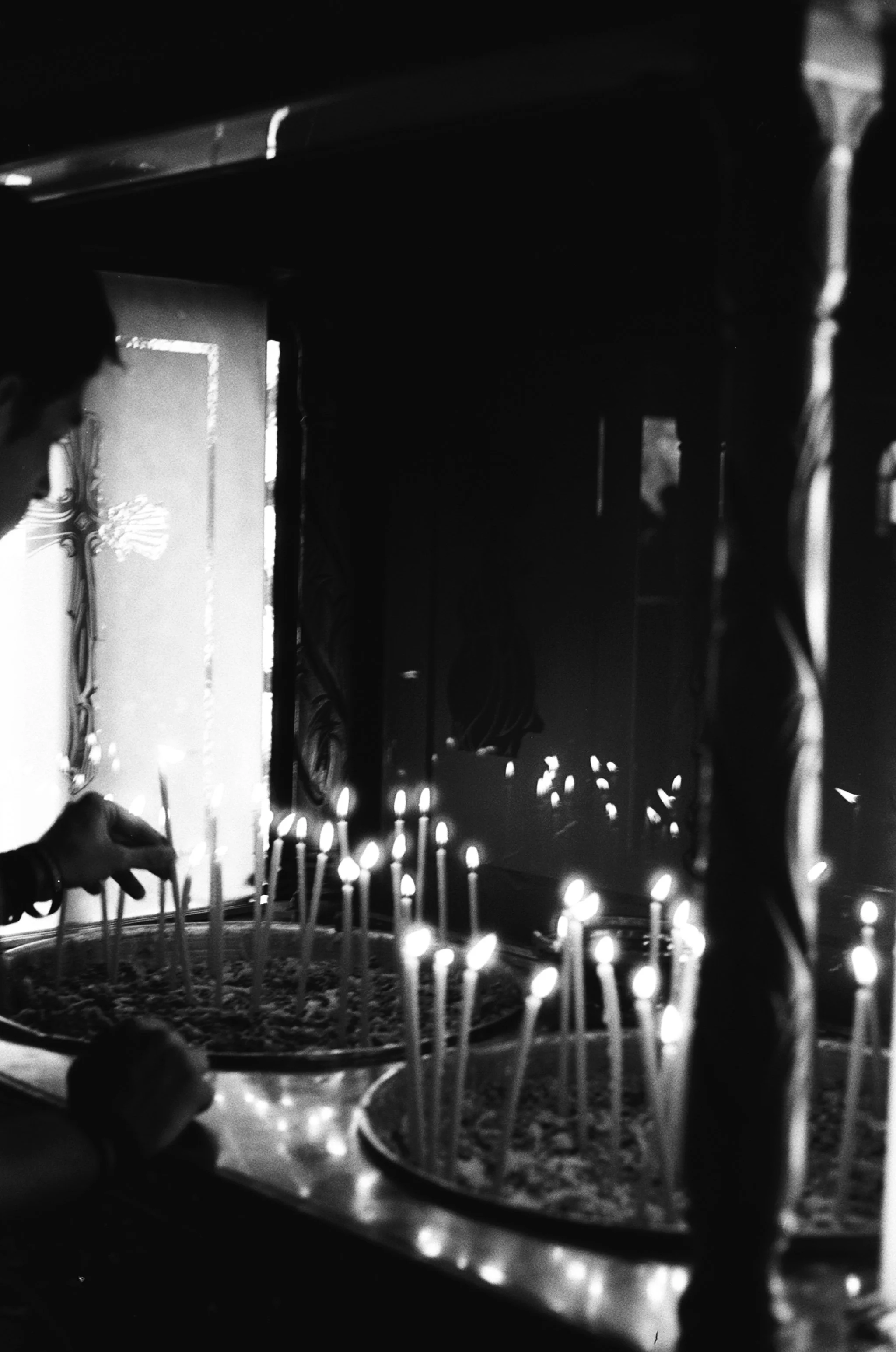 Person lighting candles at a religious shrine, with illuminated candles on a table and a framed religious picture or icon in the background.