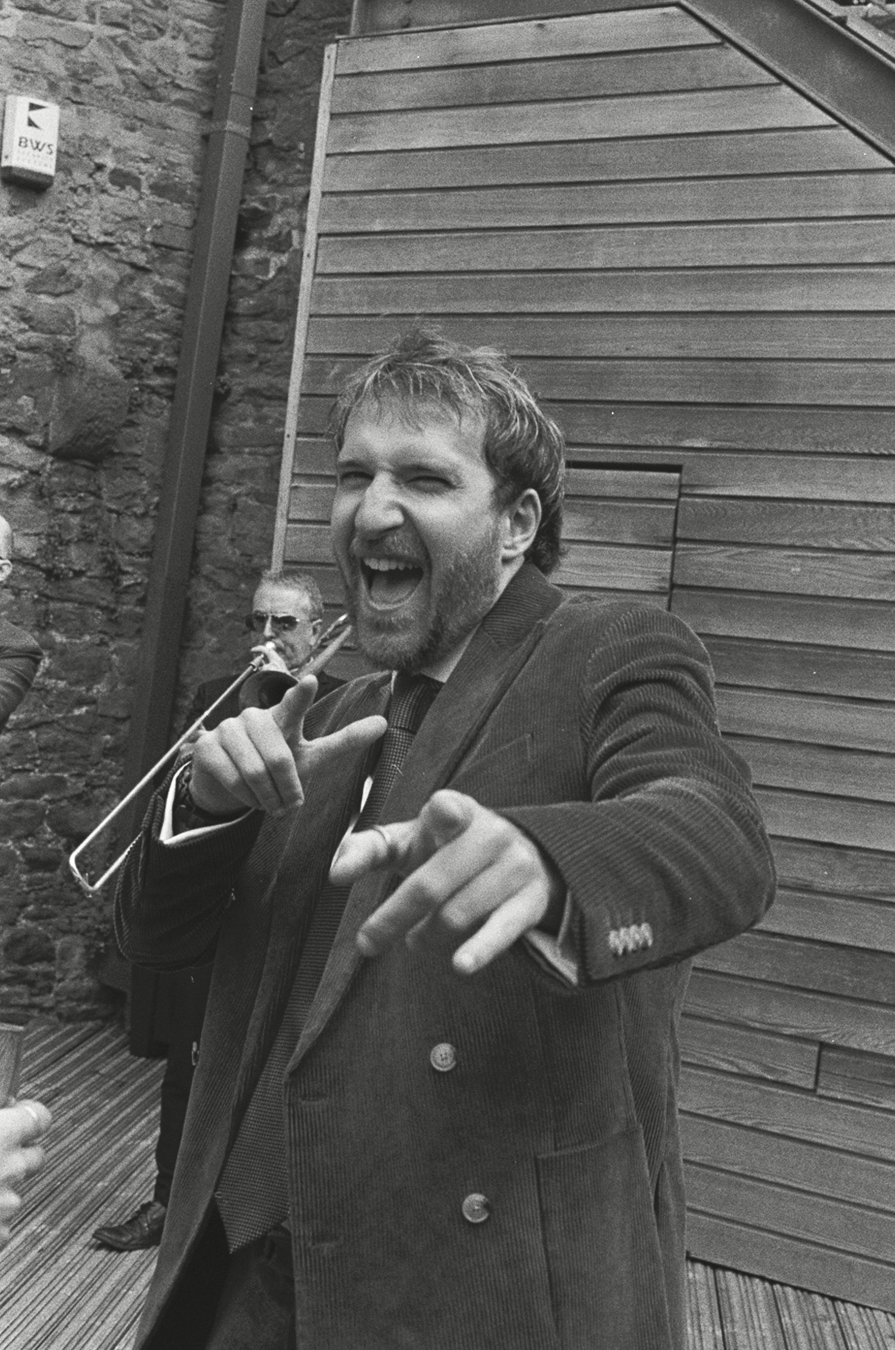 A man with a beard in a suit smiling and pointing at the camera, with another man in sunglasses in the background playing a trombone, against a stone wall and wooden paneling.