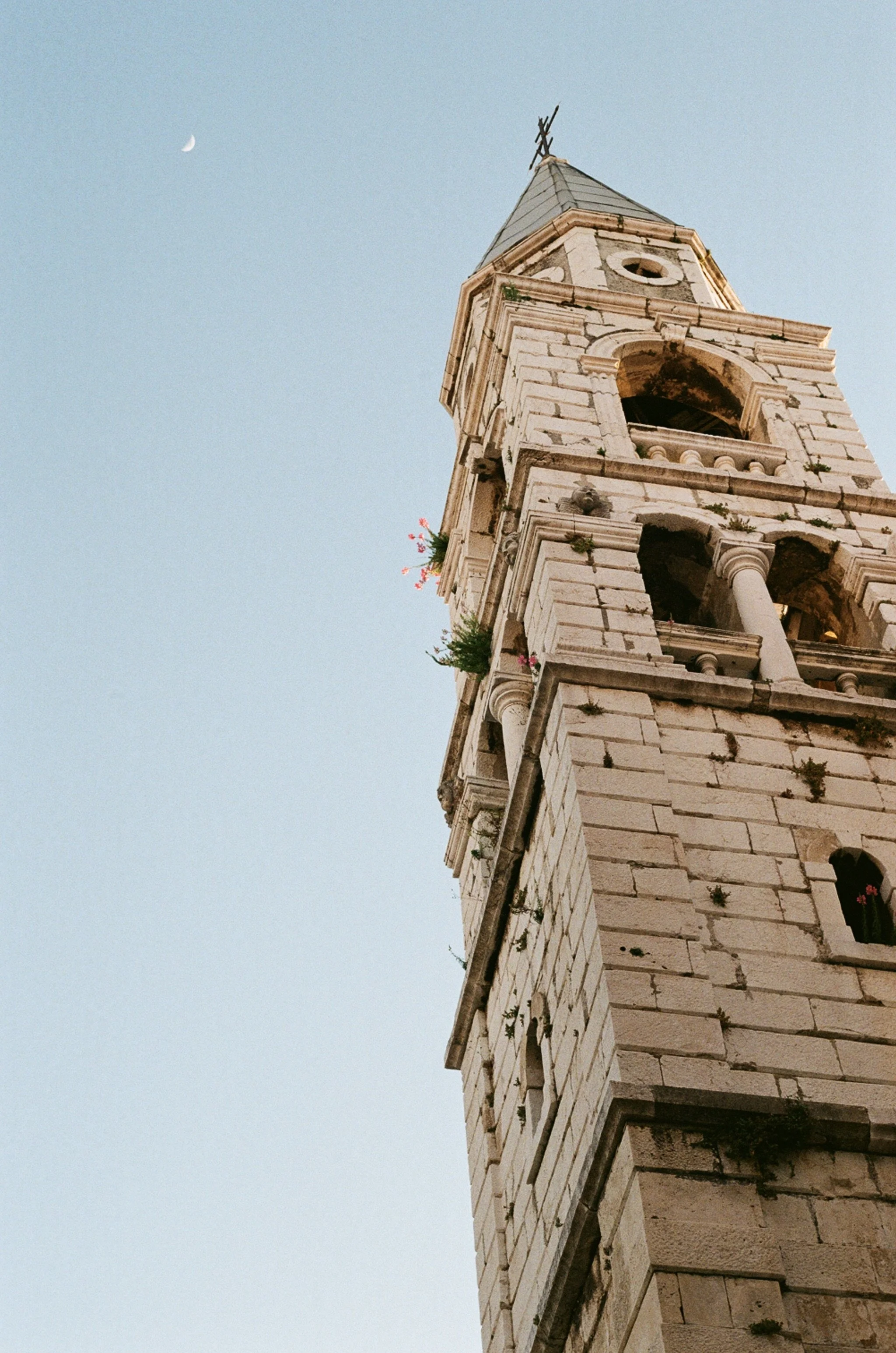 A tall, ancient stone clock tower with archaeological details and small plants growing in its crevices, against a light blue sky with a faint crescent moon visible.