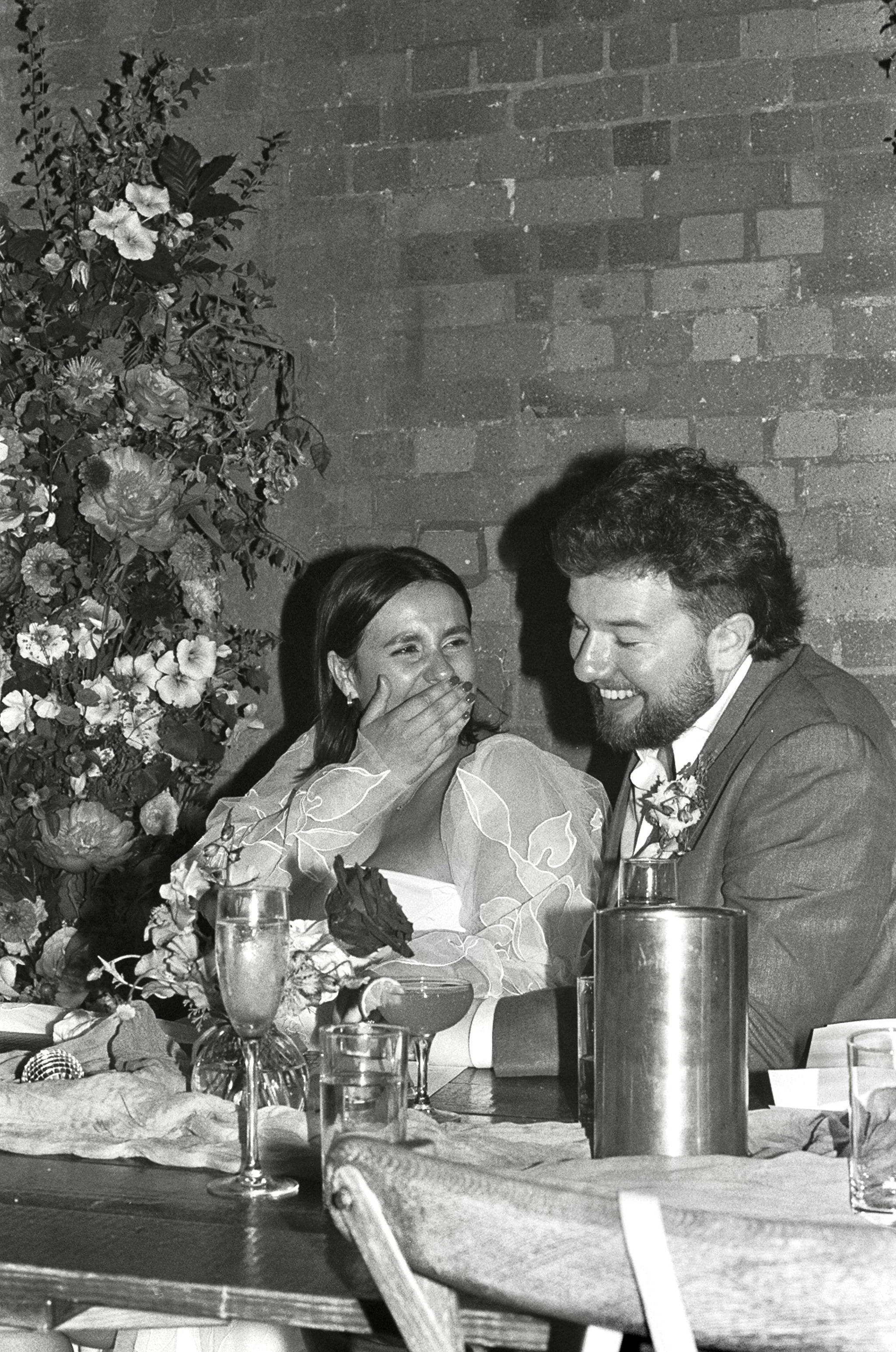 A black and white photo of a woman and a man at a wedding reception, sitting at a table with drinks, flowers, and a brick wall in the background. The woman is covering her mouth while smiling, and the man is smiling back.