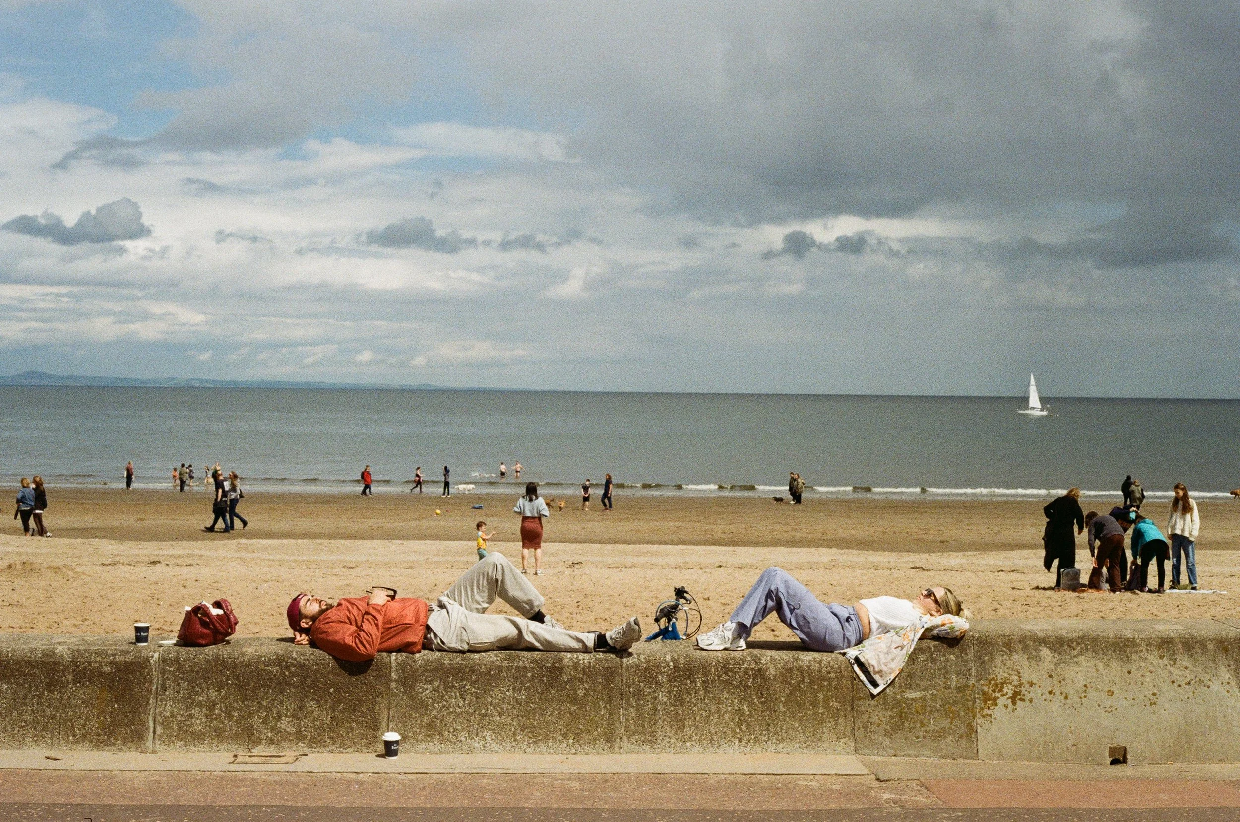 People relaxing on a concrete barrier by the beach with others walking and enjoying the shoreline. There is a sailboat on the water under a cloudy sky.