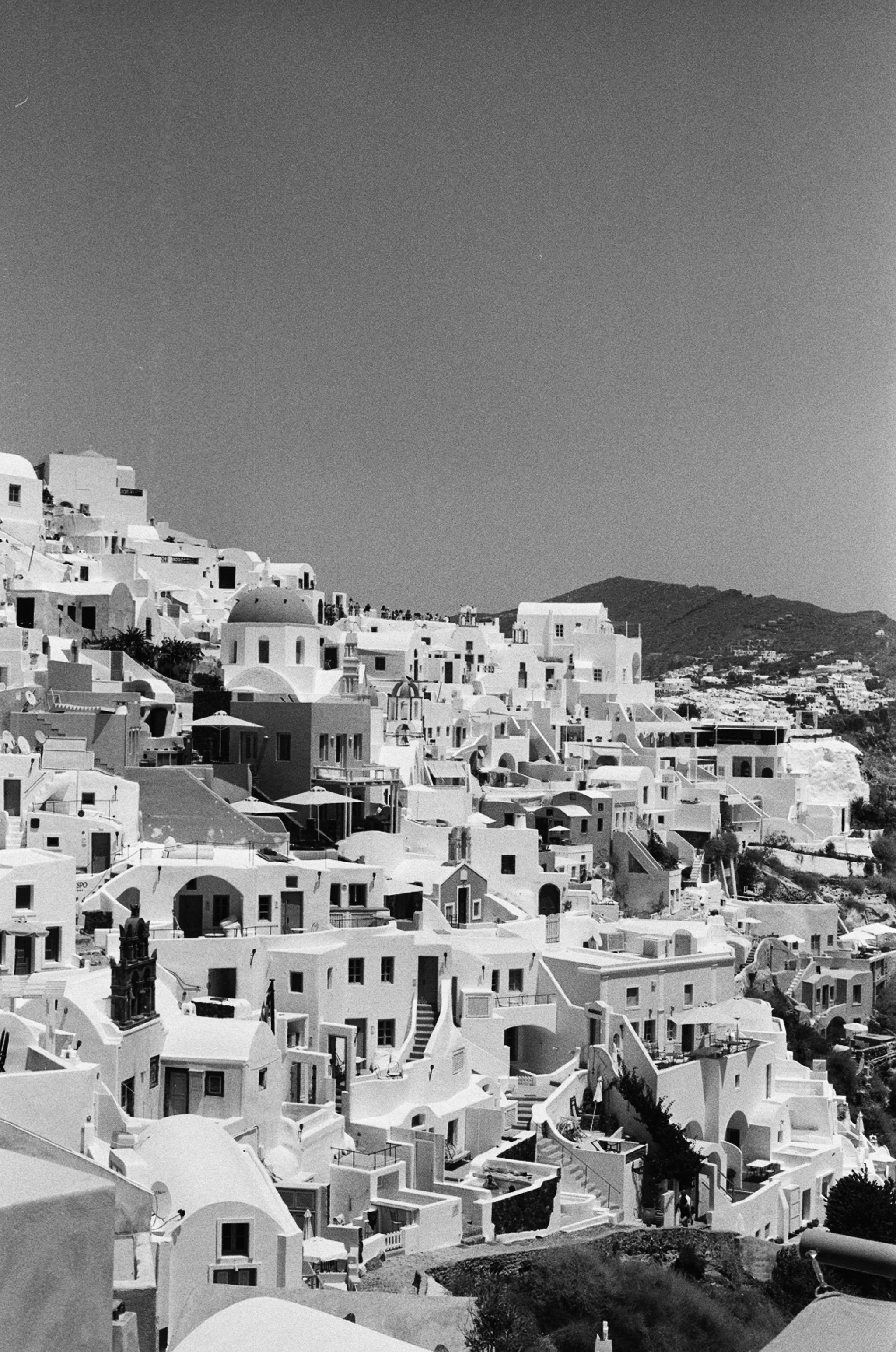 A black and white photo of white buildings on a hillside, typical of a Greek island village, with a mountain in the background.